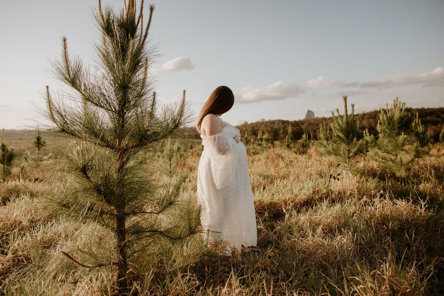 Woman in white dress standing among young pine trees in an open field under a partly cloudy sky.