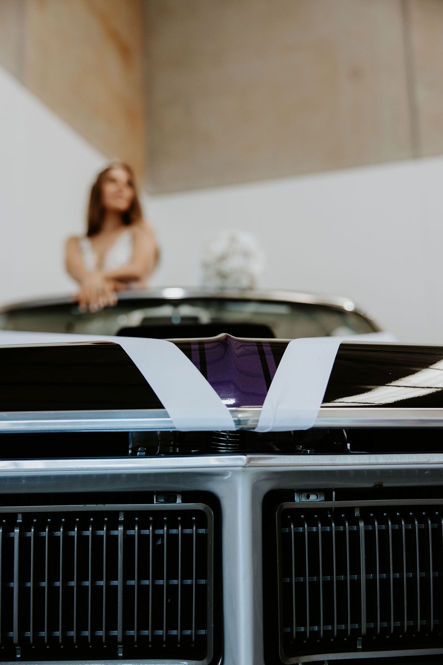 Close-up of a vintage car hood with white ribbon, blurred bride in white dress standing behind it.