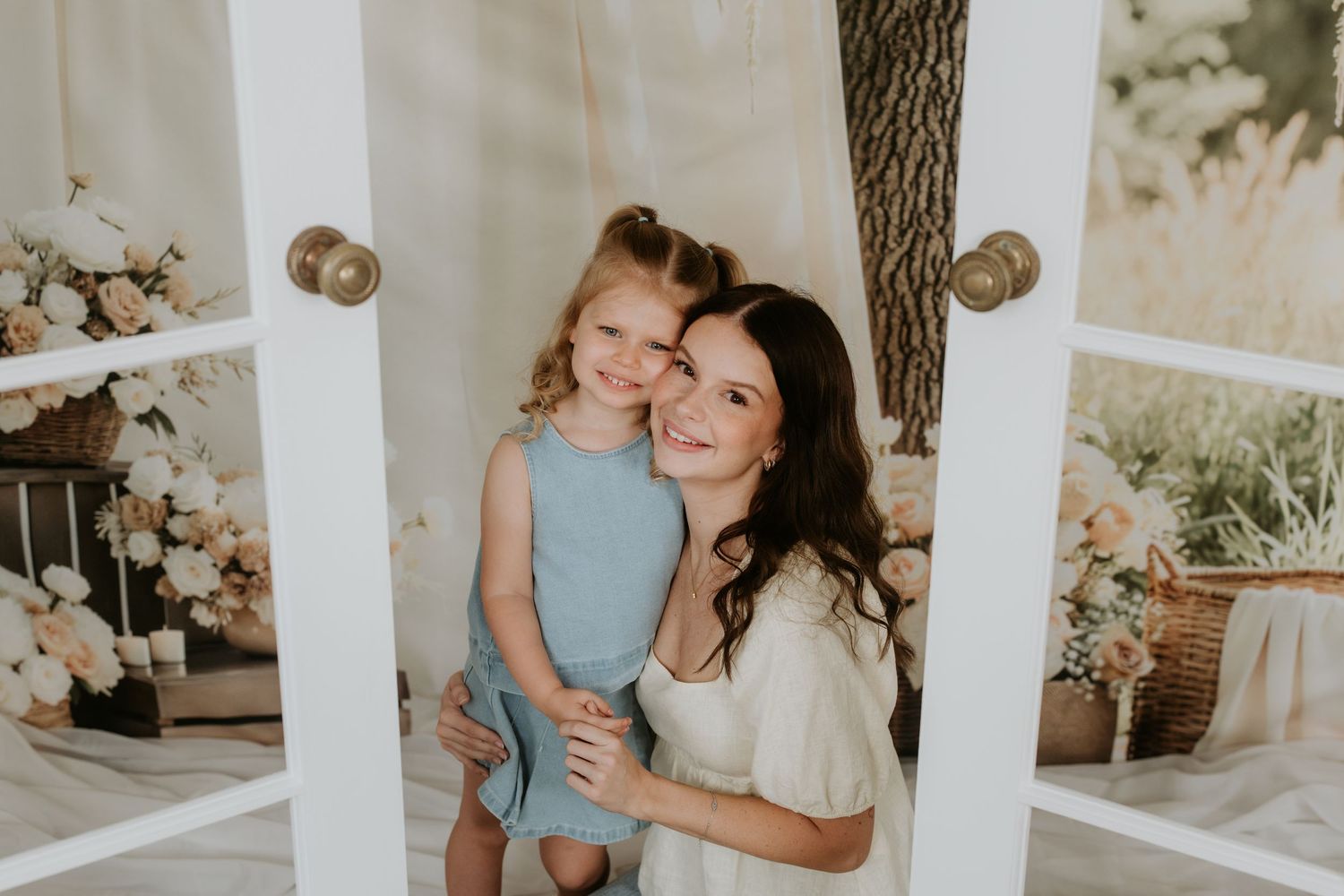 A smiling mother and young daughter in a blue dress embrace in a white doorframe with floral decor.