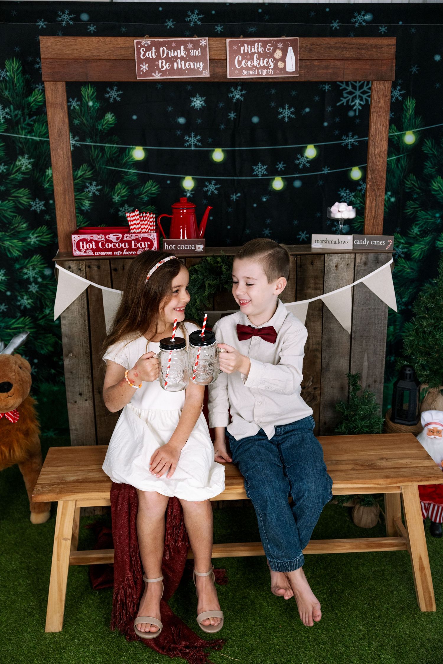 Children share hot cocoa at Christmas-themed photo booth with string lights and wooden backdrop.