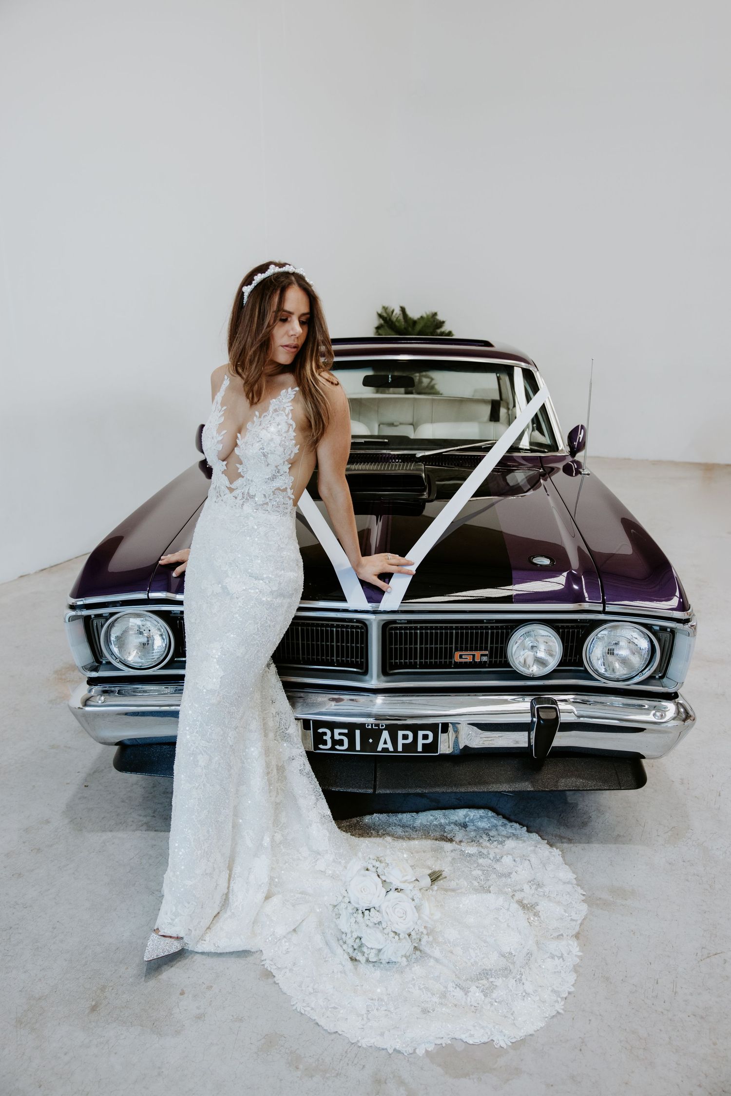 Bride in lace wedding gown with train poses beside a vintage purple muscle car decorated with white ribbons.