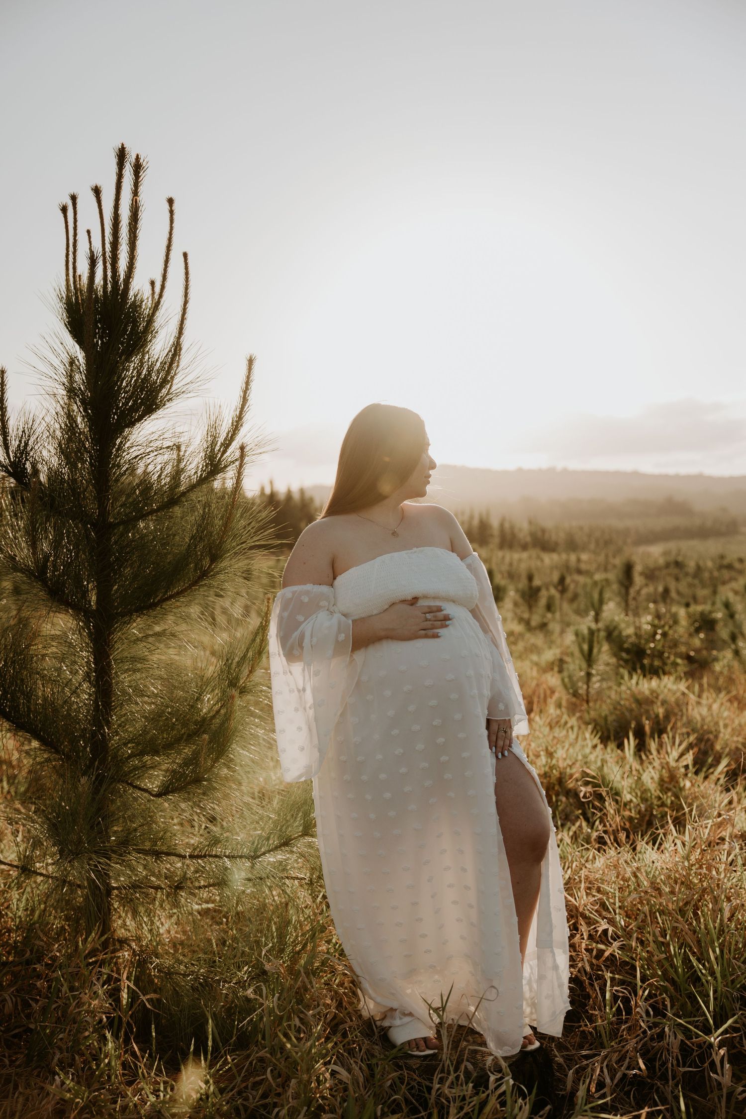 Woman in white dress poses near pine tree during golden hour.