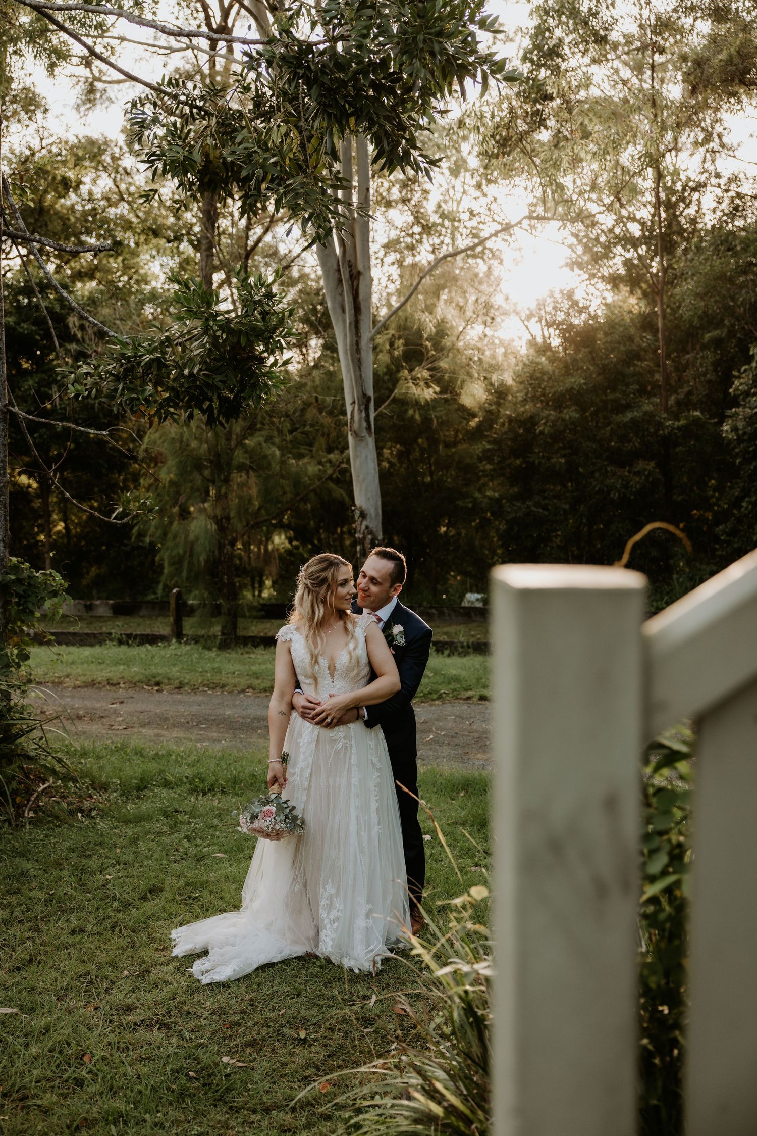 Wedding couple shares an intimate moment in a garden setting with natural lighting and white fencing.