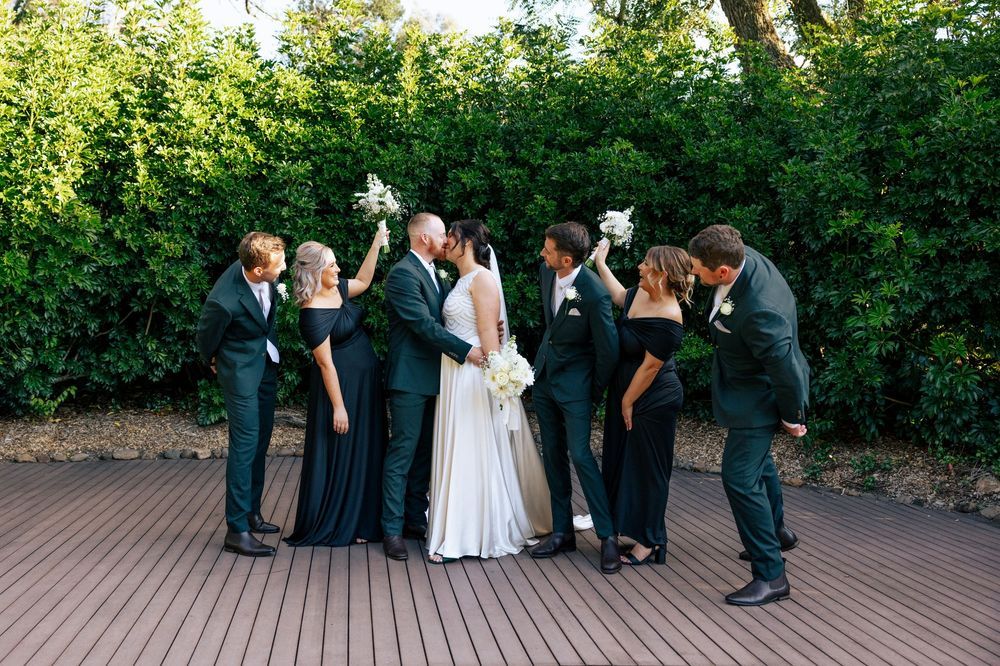 Wedding party poses for formal group photos on outdoor deck surrounded by greenery.