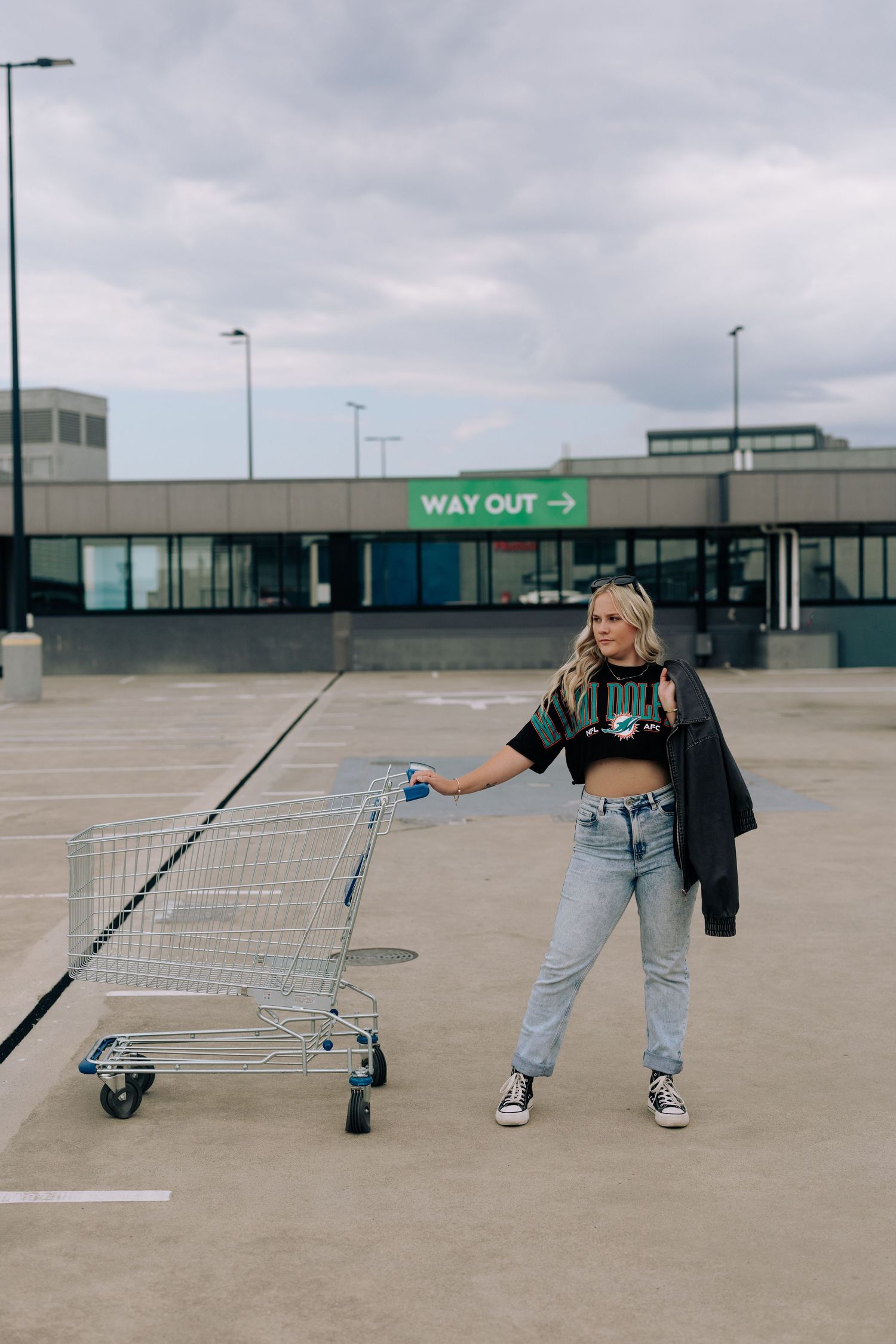 Person in crop top and jeans posing with shopping cart in empty parking lot.