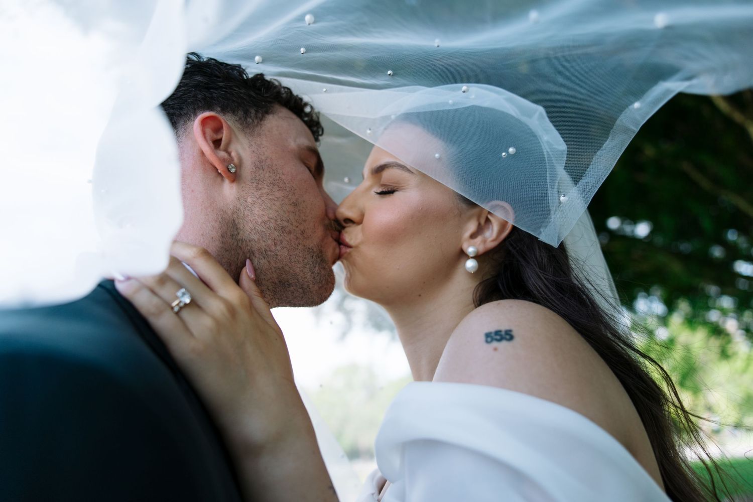 Bride and groom share a romantic kiss under a flowing white veil outdoors on their wedding day.