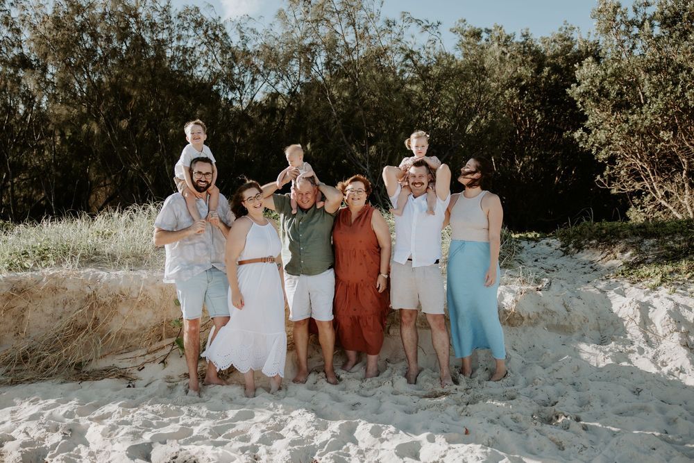 Casual group portrait session on a sandy beach with natural coastal vegetation in the background.