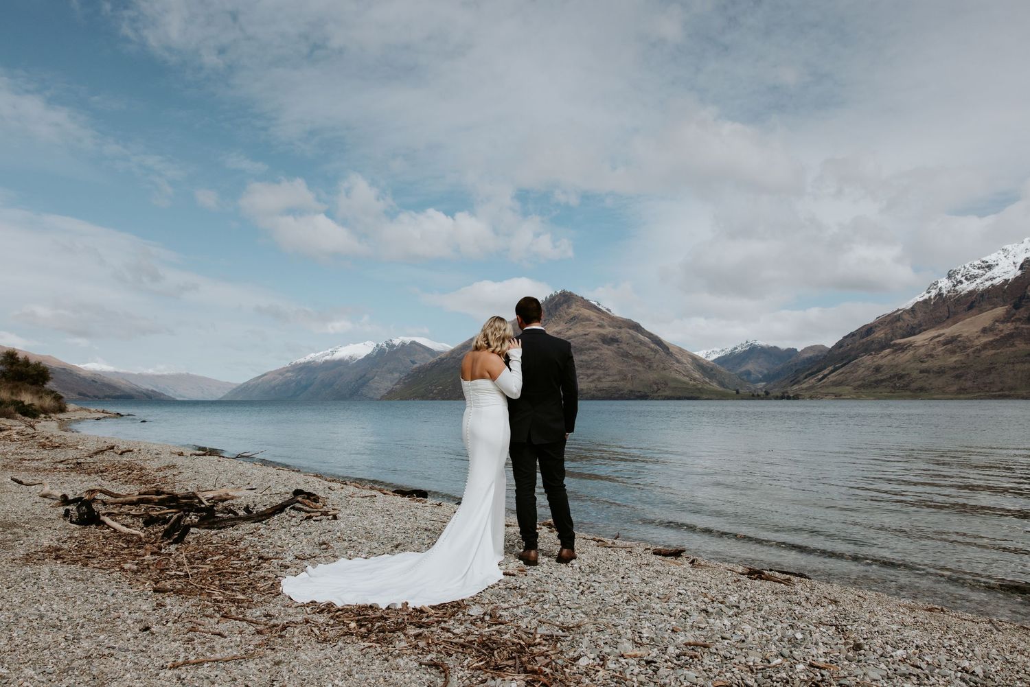 Romantic wedding photo by mountain lake with cloudy sky backdrop.