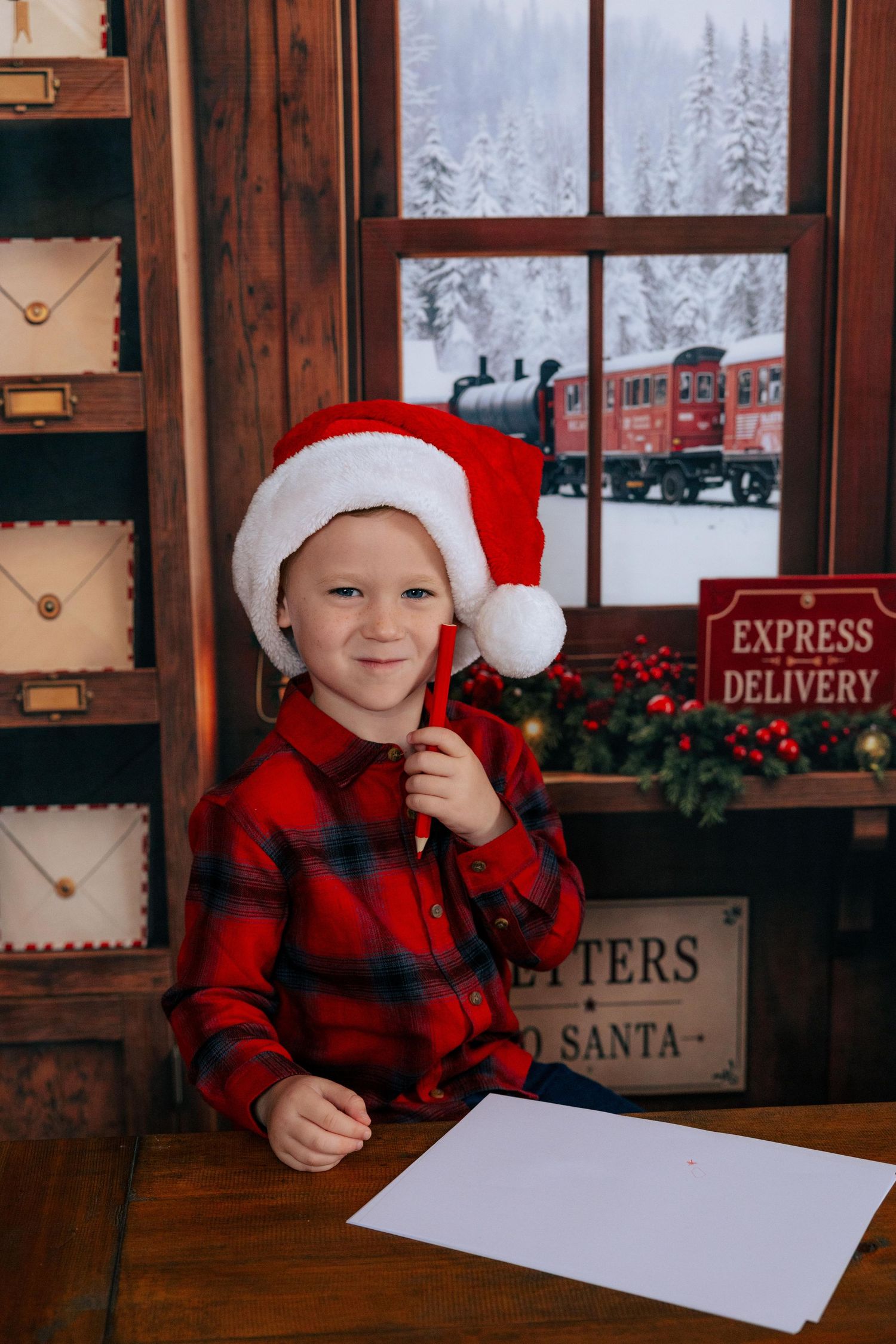 A cheerful boy in a Santa hat and red plaid shirt writes a letter to Santa at a festive Christmas post office desk.