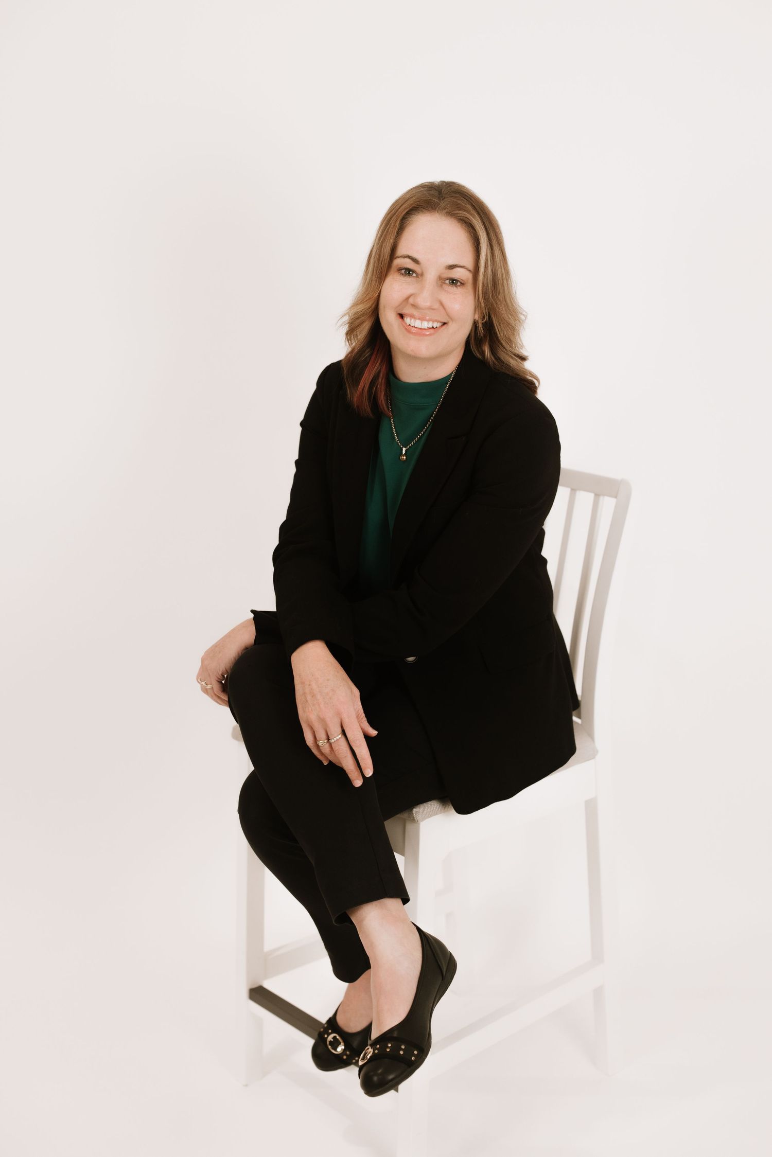 Business professional poses on white chair wearing black suit and green top.
