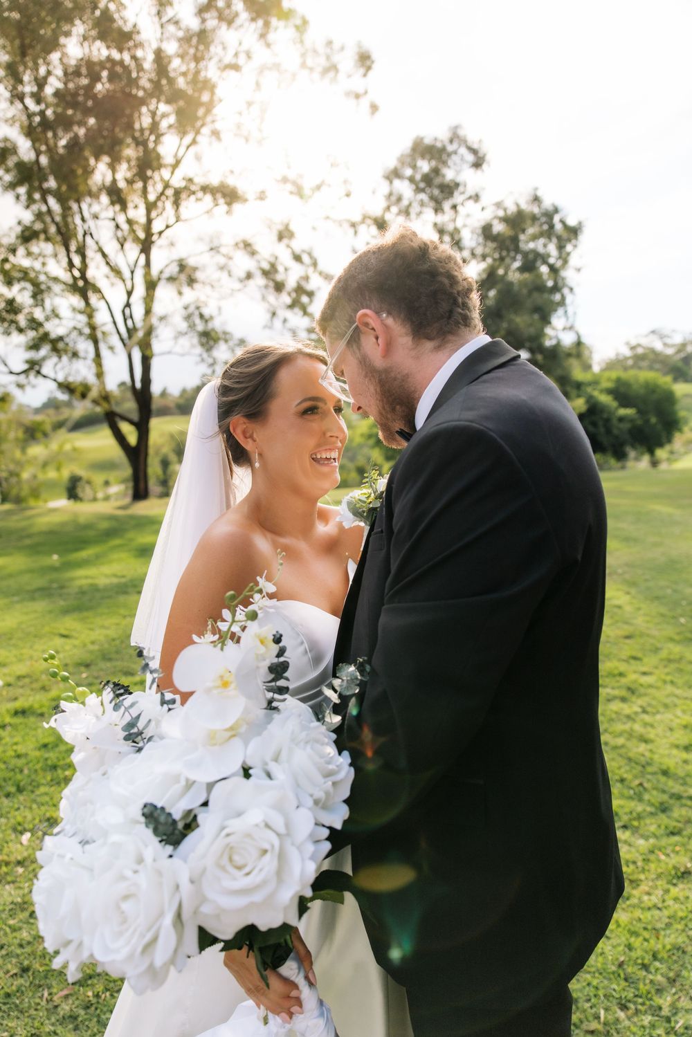 Wedding couple poses with white roses in a sunlit garden setting.