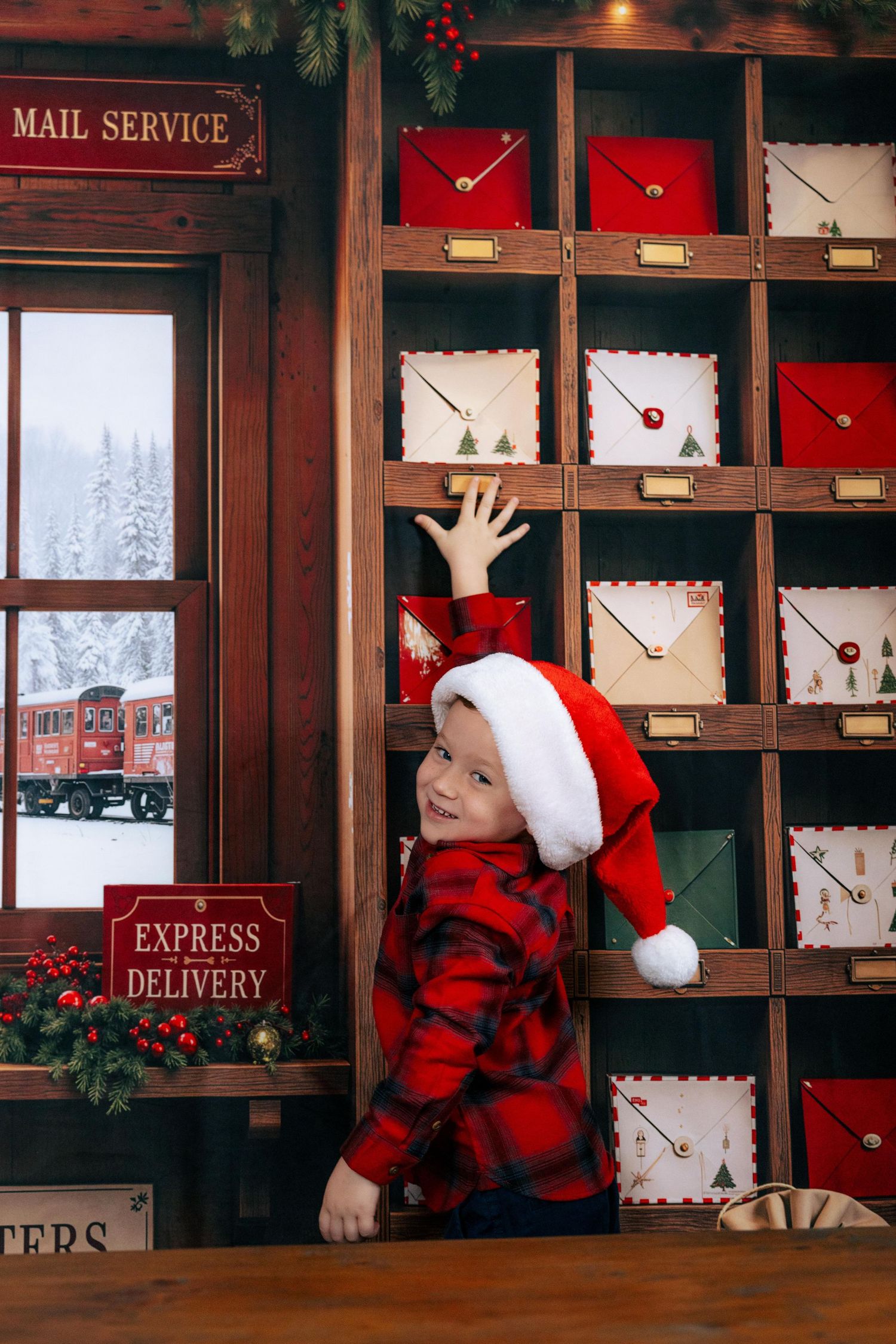 A cheerful boy in a Santa hat and plaid shirt reaches for Christmas mail in a festive holiday post office setting.