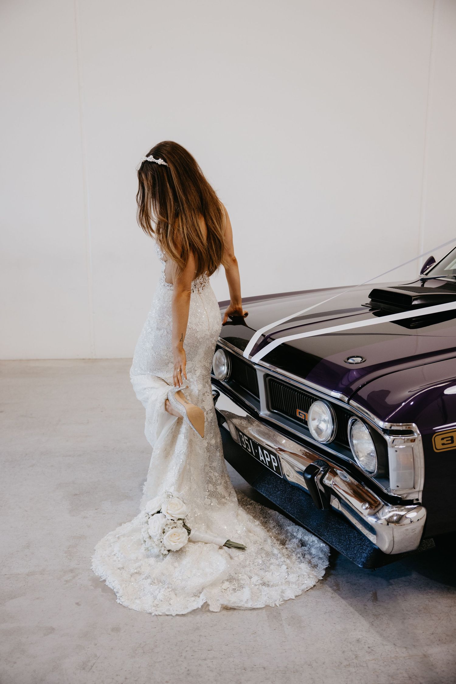Bride in lace wedding gown with train stands beside a vintage purple classic car holding a white bouquet.