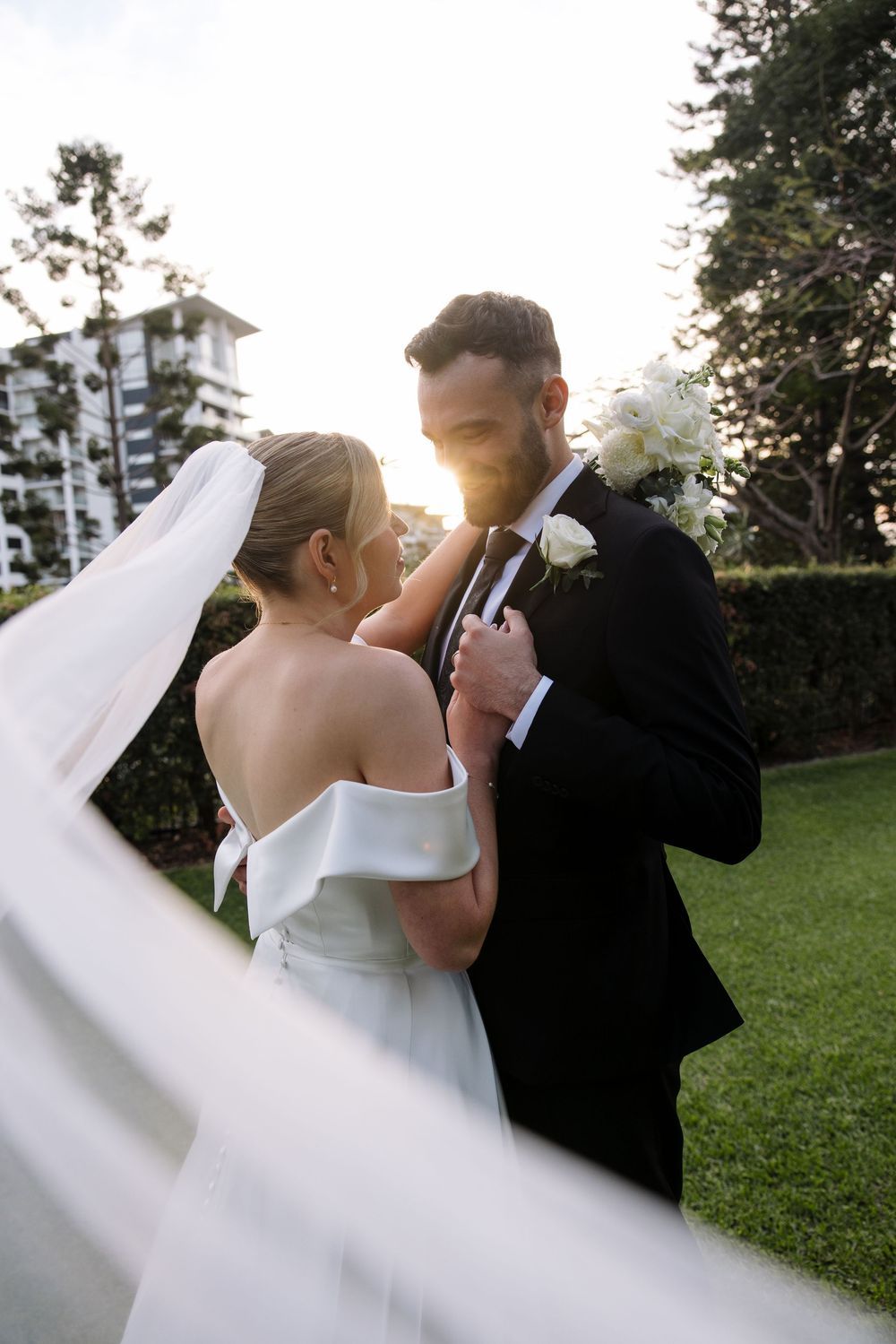 Bride and groom embrace during sunset with flowing white veil in motion.