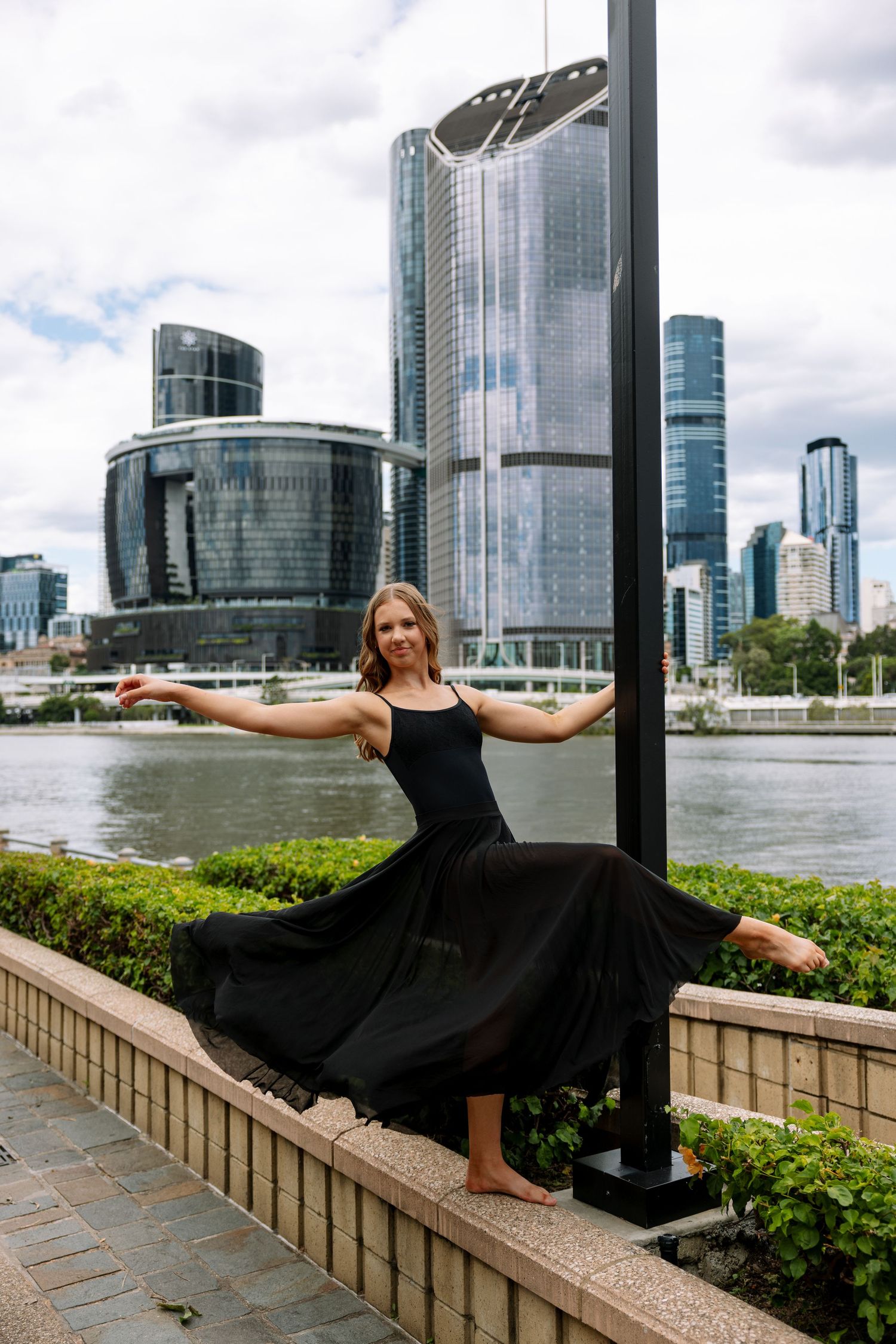 Dancer in flowing black dress performs split pose against Brisbane city skyline.