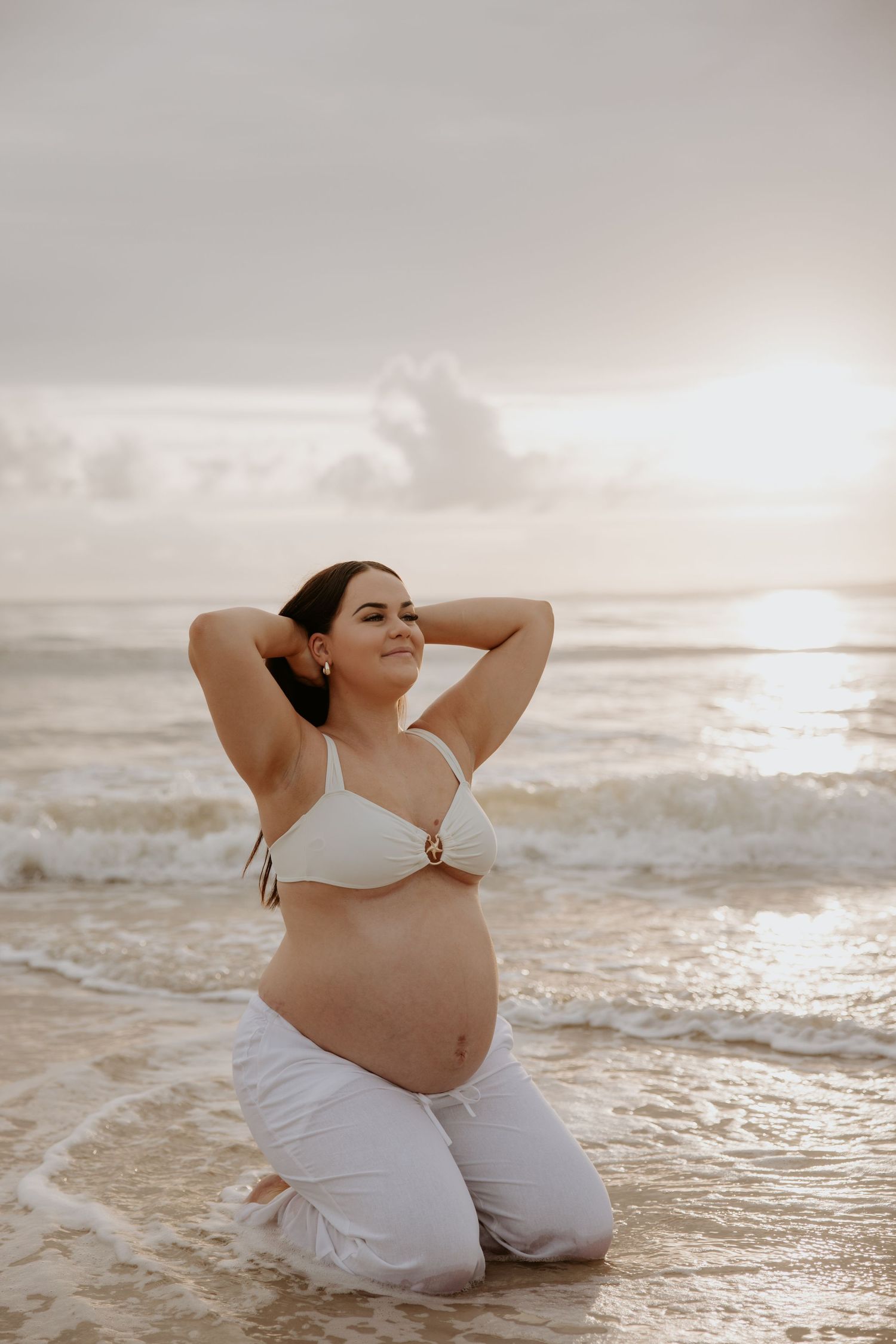 Expectant mother in white swimwear poses serenely at sunset on a beach.