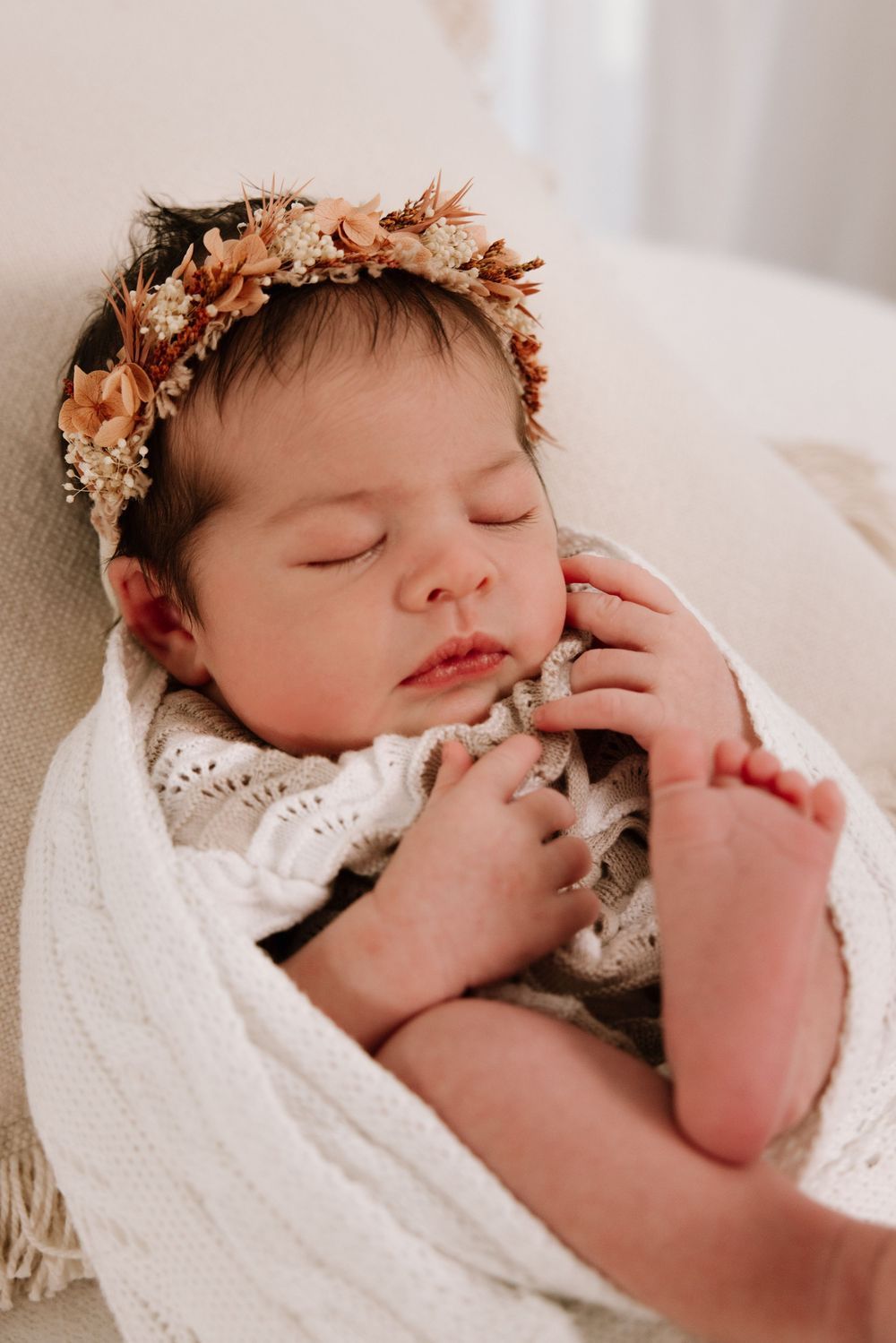 Sleeping newborn wearing delicate floral crown on white blanket.
