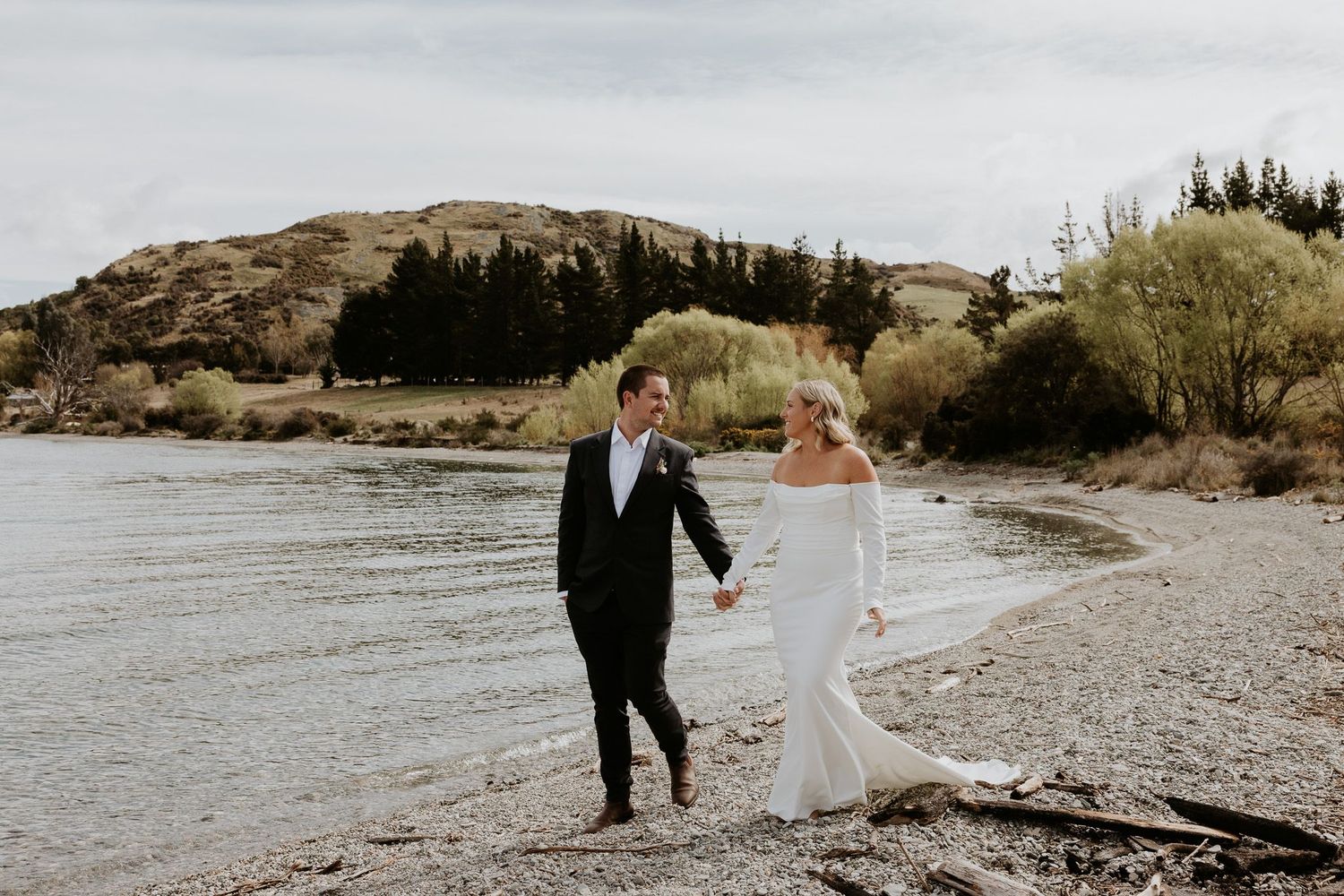 Couple walks along rocky beach shoreline with mountains and water in background.