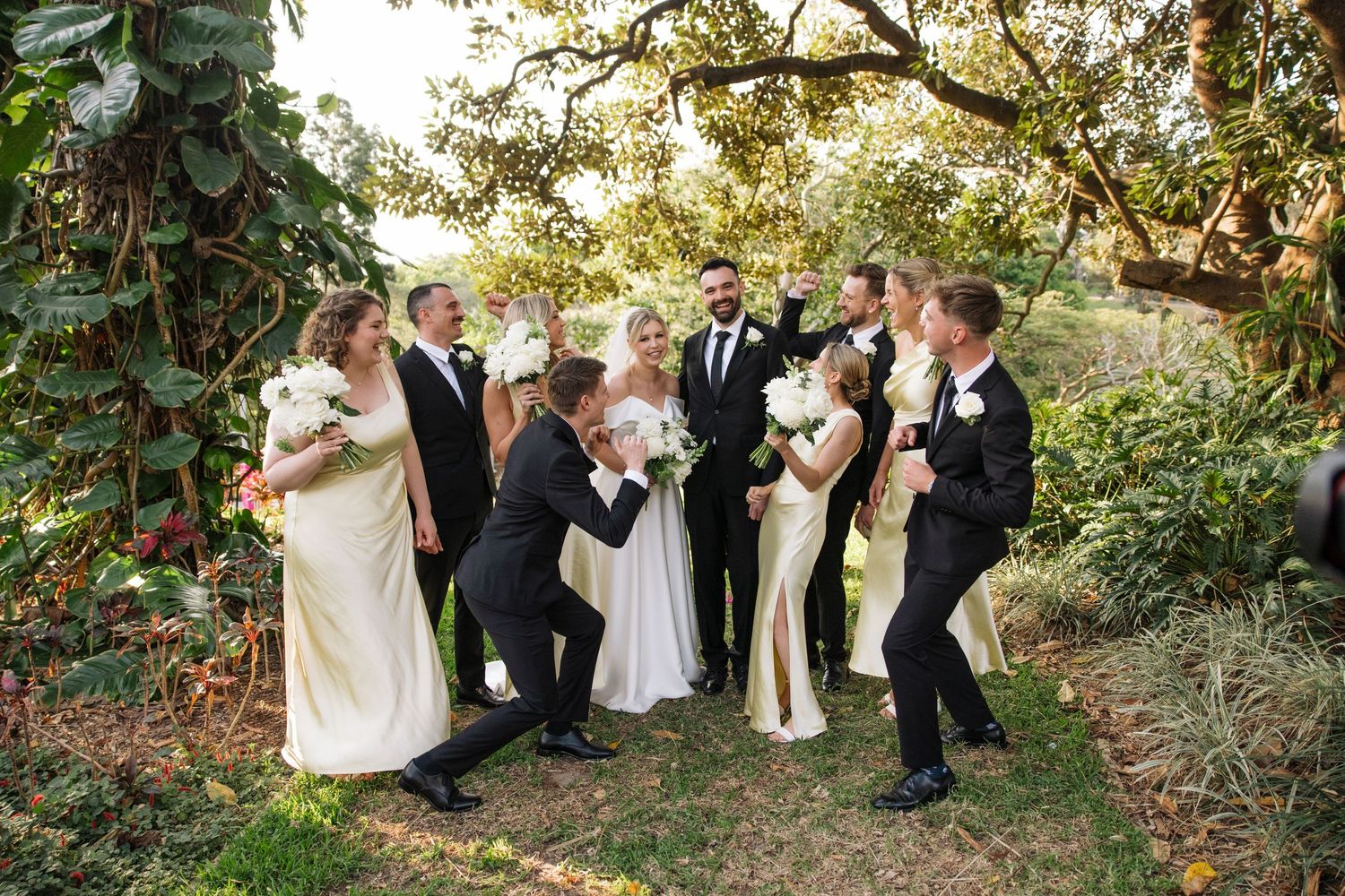 Wedding party gathered outdoors under trees for group photos during garden ceremony.