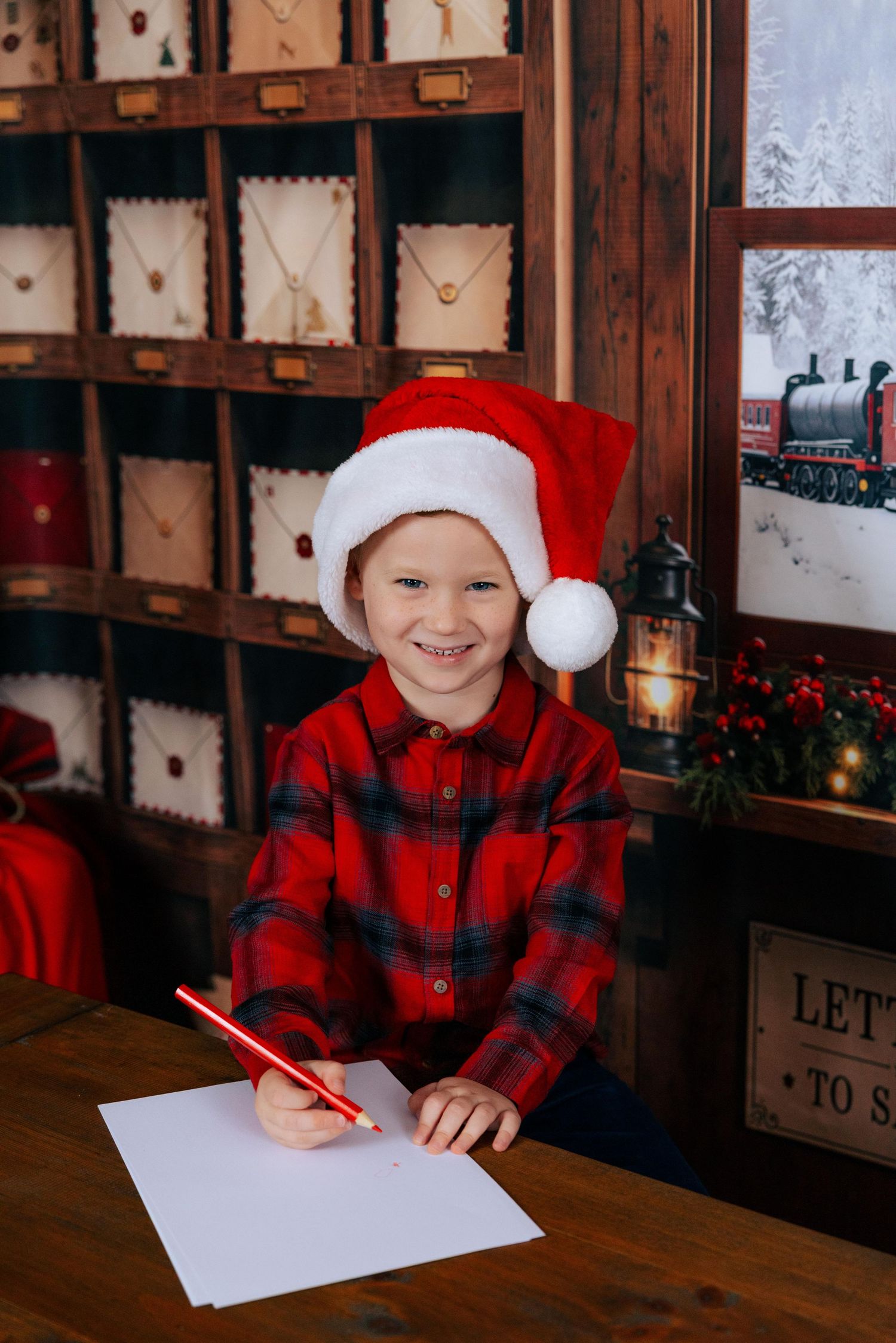 Smiling child in Santa hat and red plaid shirt writing a Christmas letter to Santa at a wooden desk.