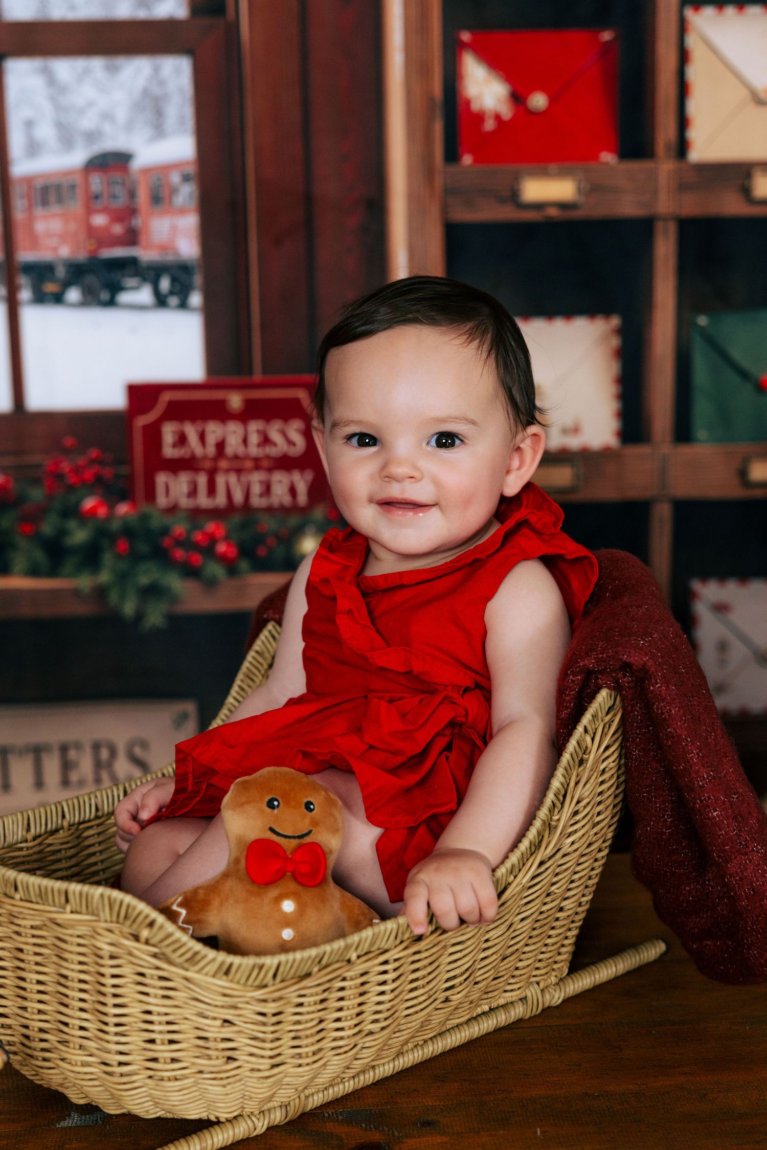 Baby in red dress sits in wicker basket with gingerbread toy, smiling at Christmas-themed photo backdrop.