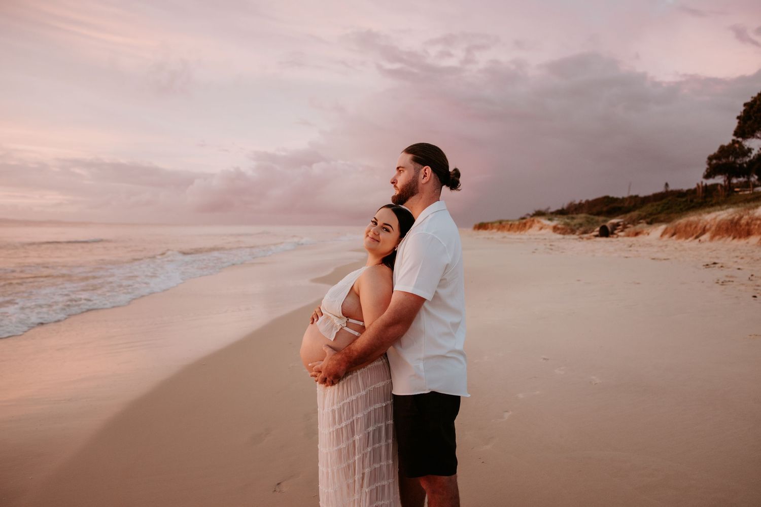 Couple sharing tender moments on a beach at sunset with pink skies.