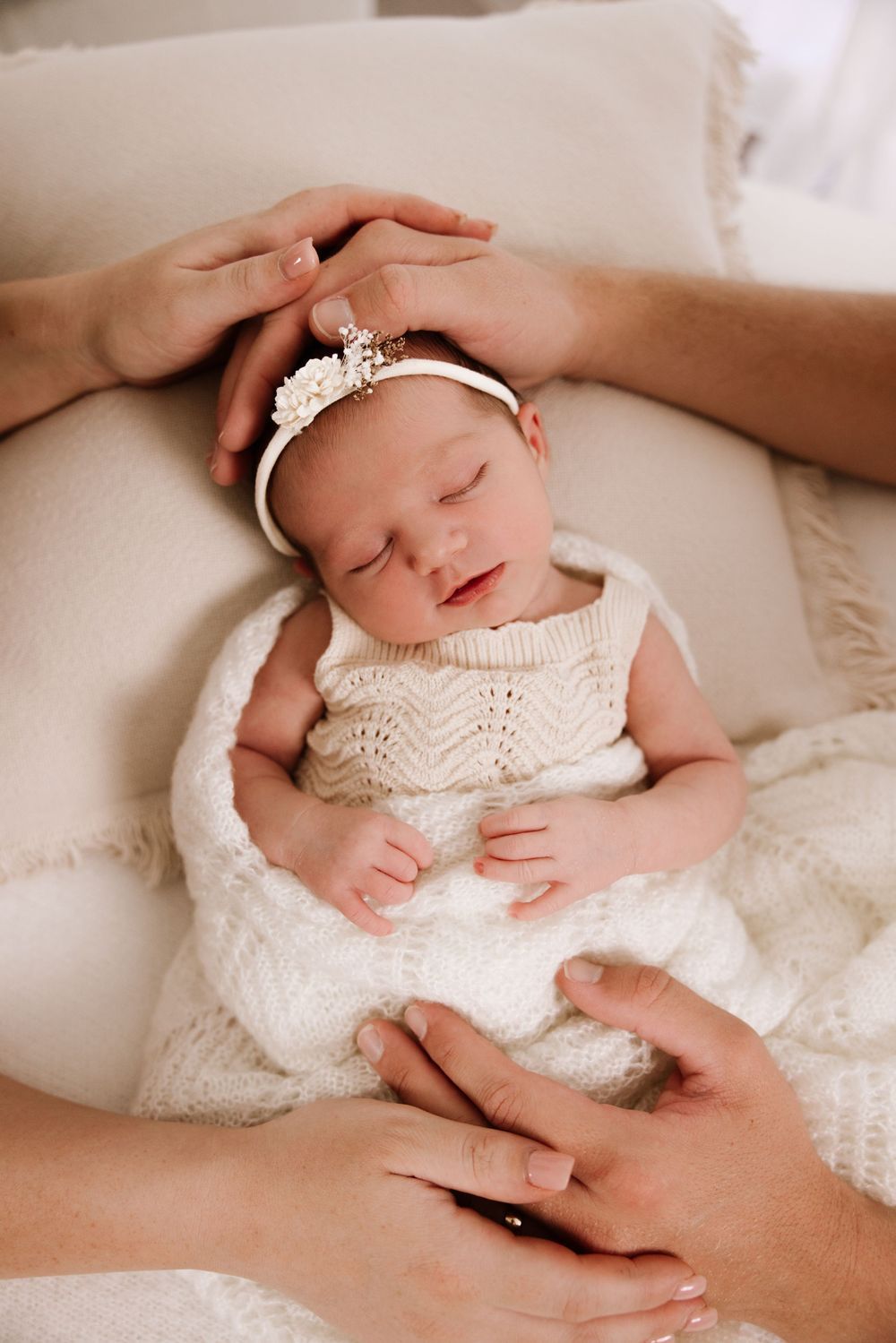 Newborn baby wearing white lace outfit lying on soft white blanket during professional photoshoot.