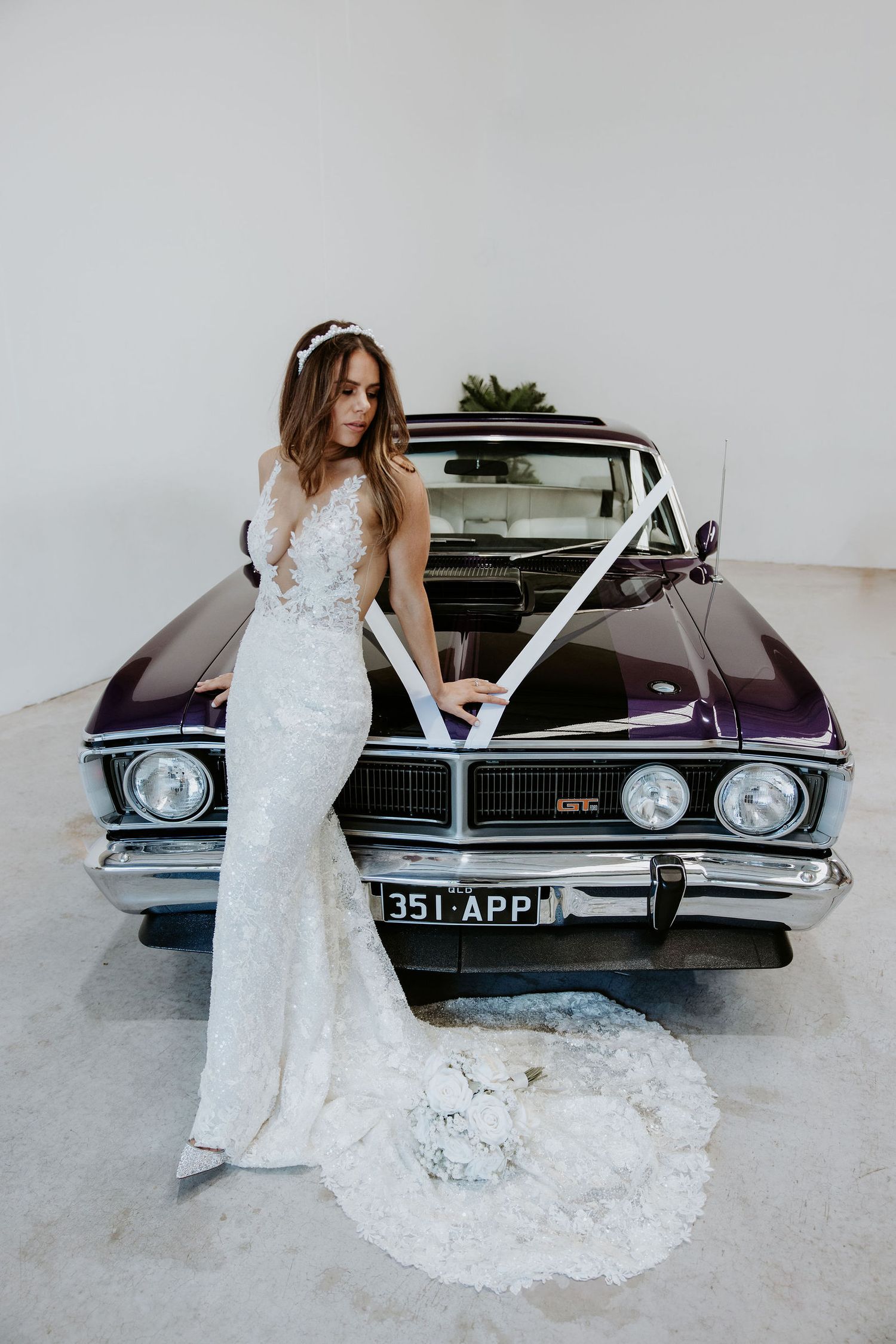 Bride in lace wedding gown poses beside a vintage purple GT car decorated with white ribbons.