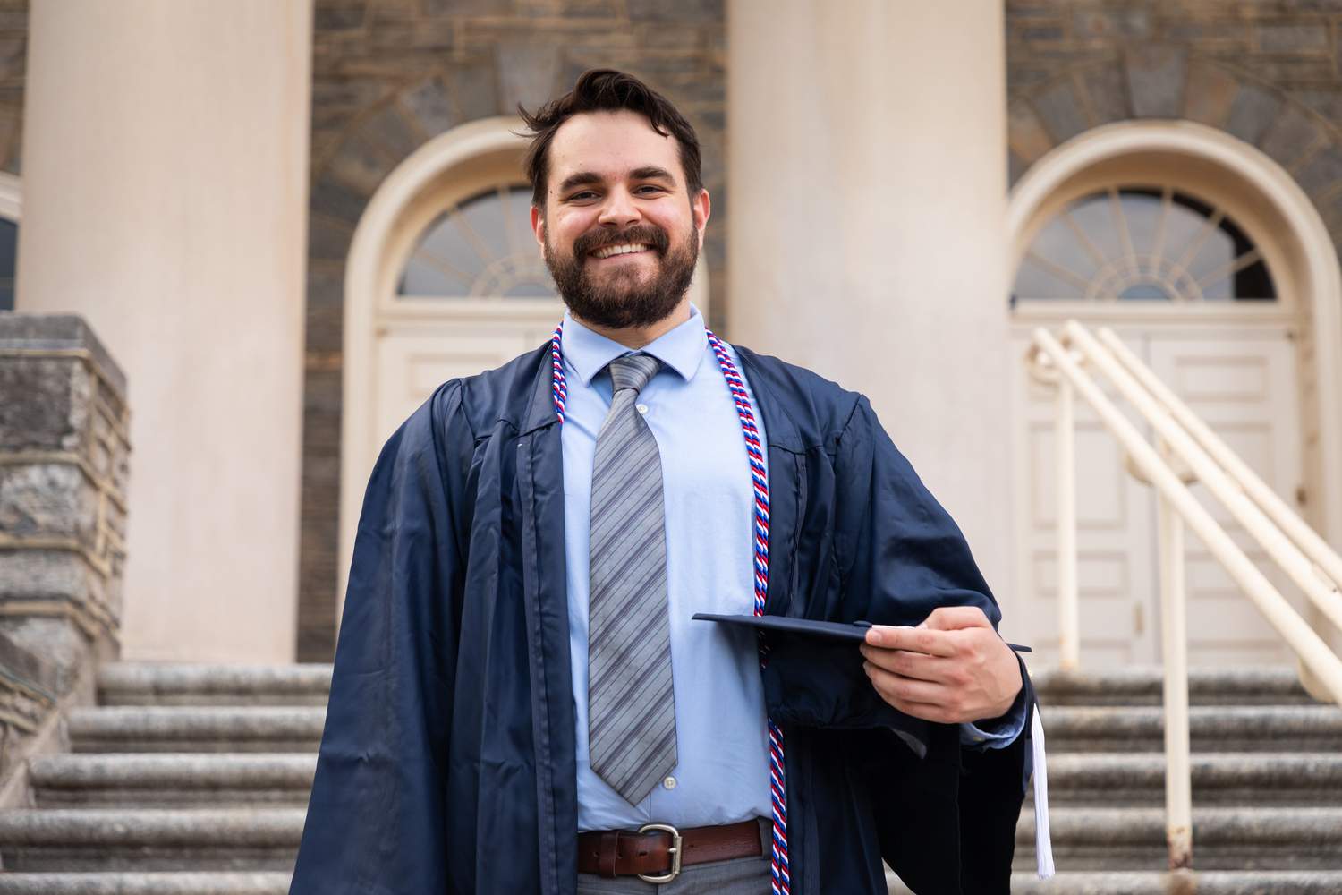 Smiling male graduate in cap and gown holding diploma outside a historic university building.