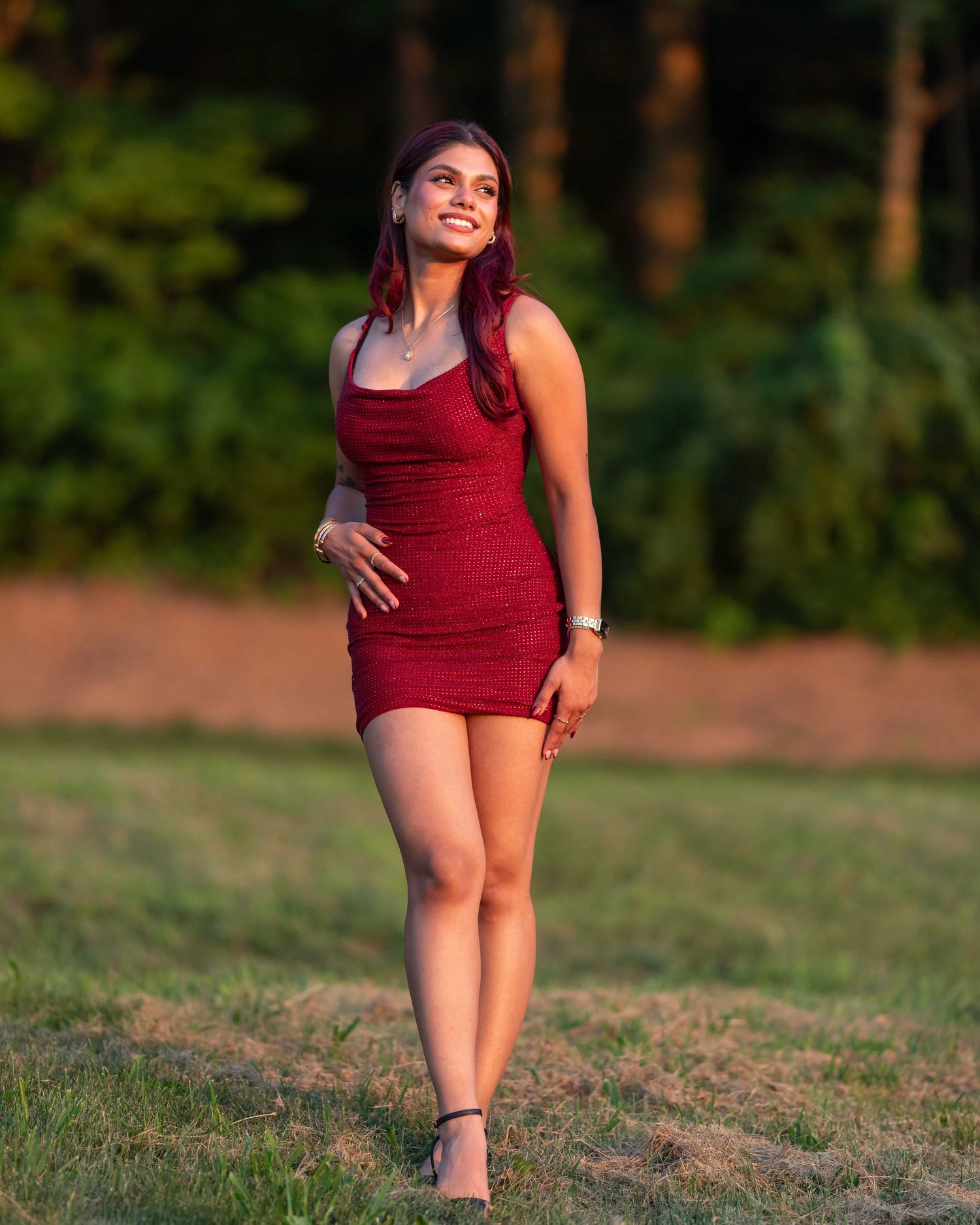 Young woman in a red mini dress walking confidently in a field during golden hour sunset.