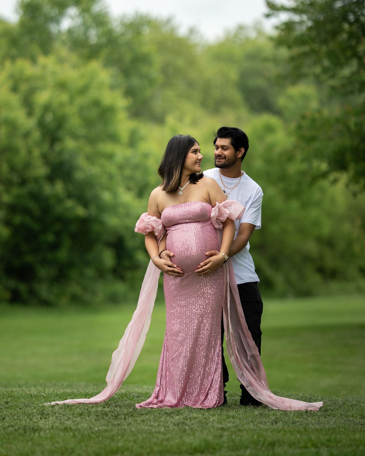 Couple posing for maternity photos outdoors, woman in pink sequin gown cradling baby bump in lush park.