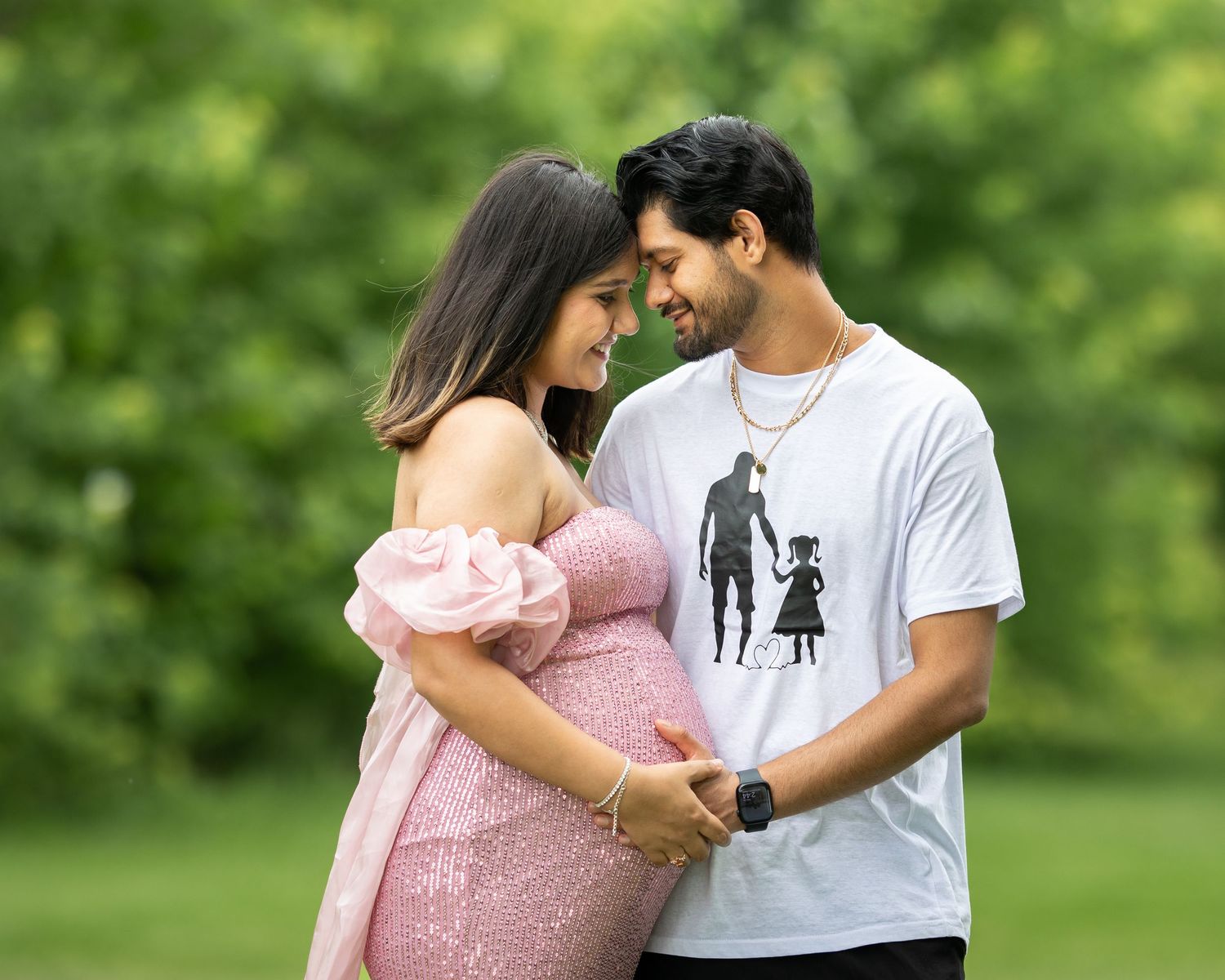 Couple sharing a tender moment outdoors during maternity photoshoot in a green park setting.