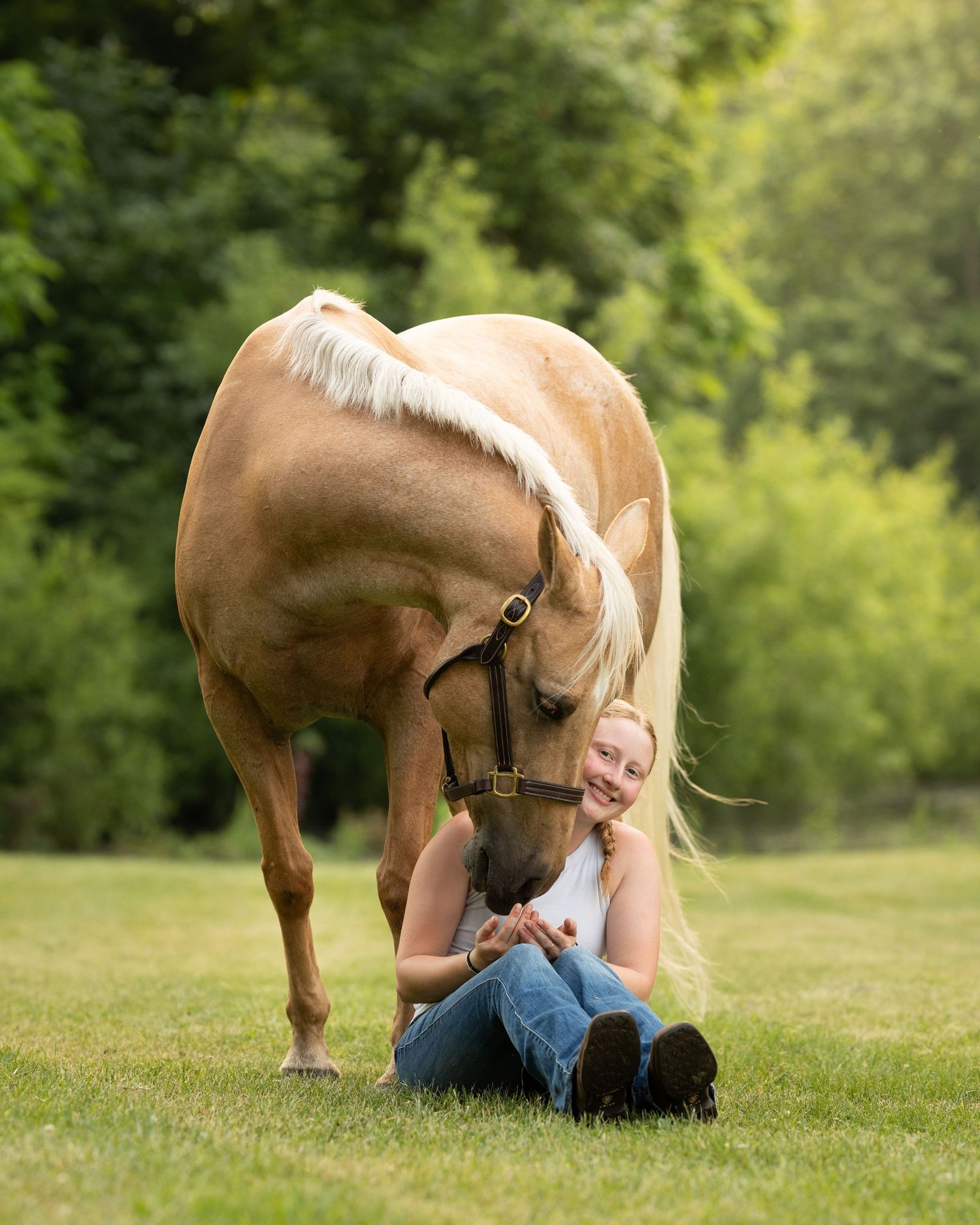 Blonde woman sitting on grass laughing as a palomino horse nuzzles her in a lush green outdoor setting.