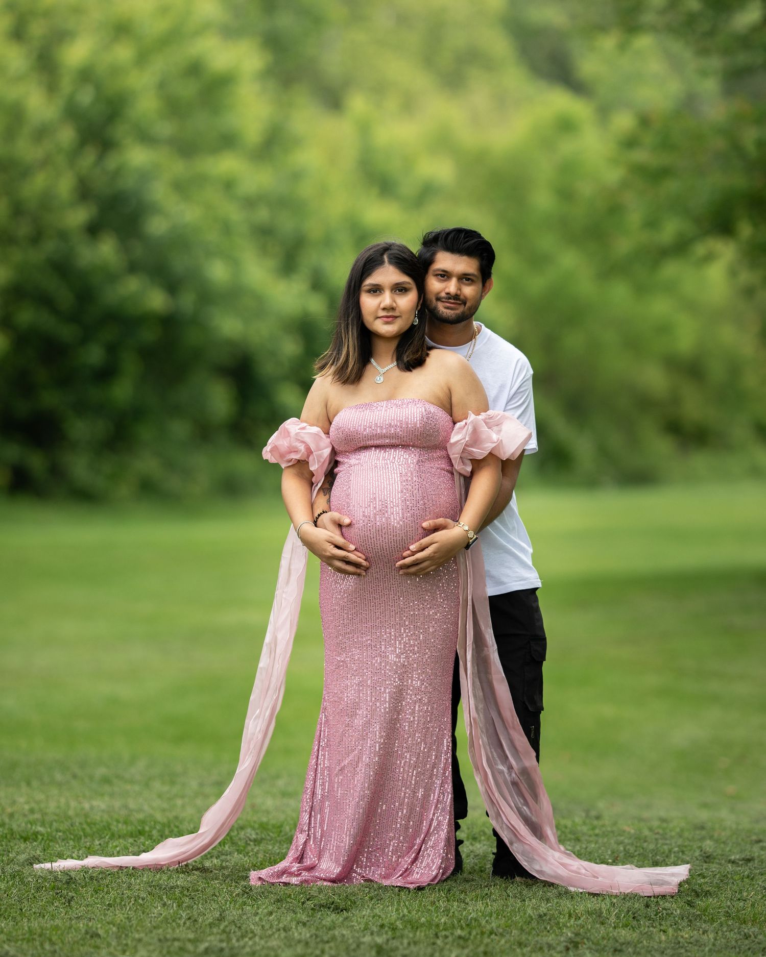 Couple posing for elegant maternity photos in a green park, woman in pink sequin gown.