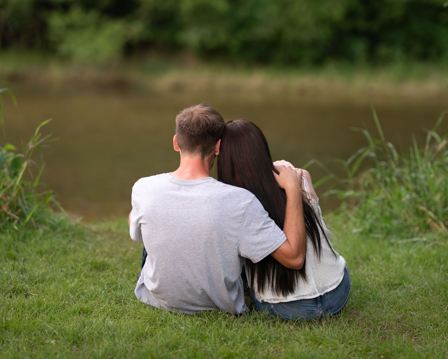 Couple sitting together by calm pond with woman leaning on mans shoulder.