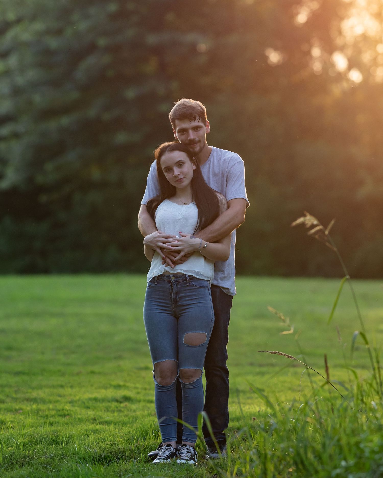 Couple embracing in golden sunset light on green grass with trees behind them.