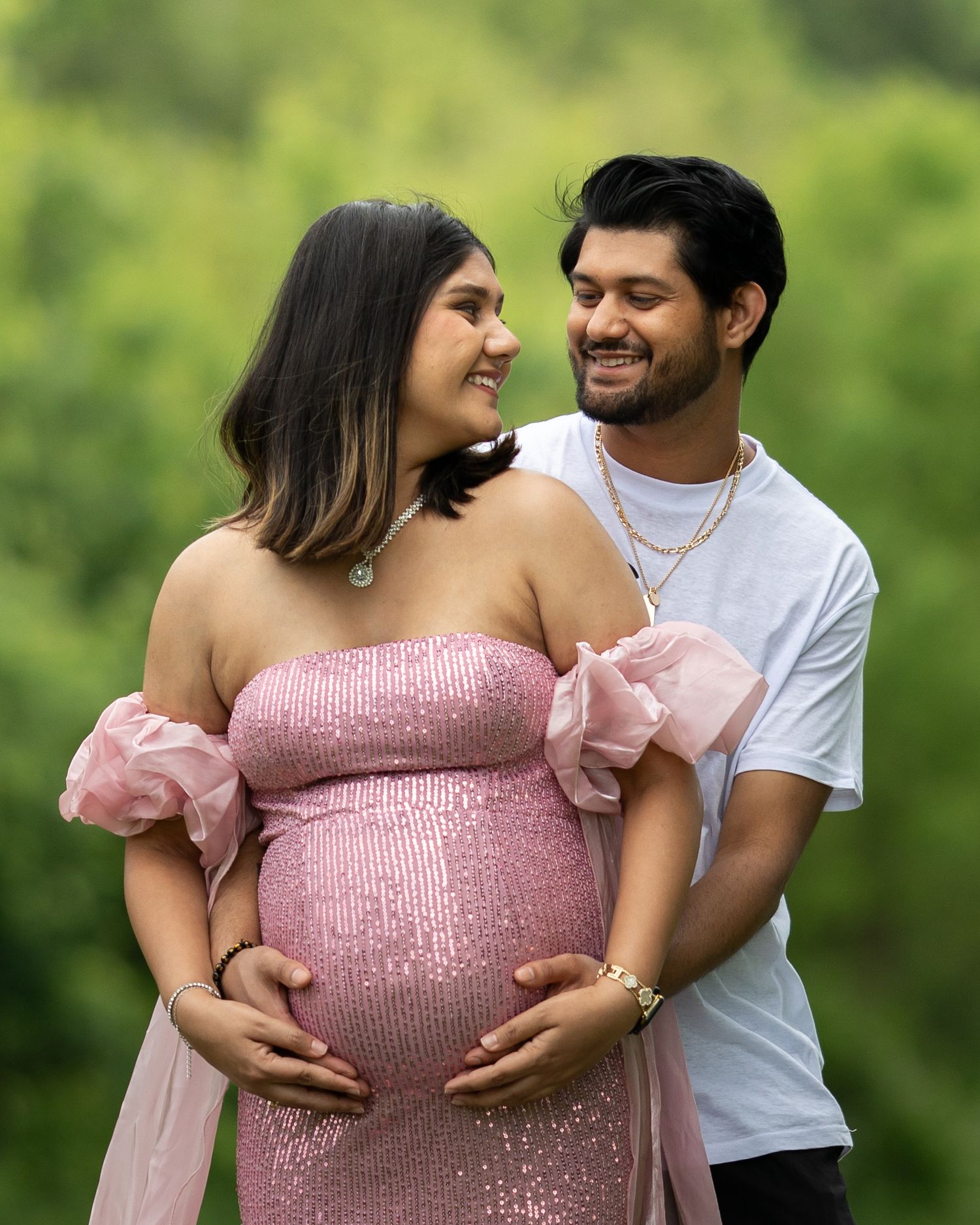 Smiling couple sharing a joyful maternity photo outdoors, woman in pink sequin dress holding baby bump.