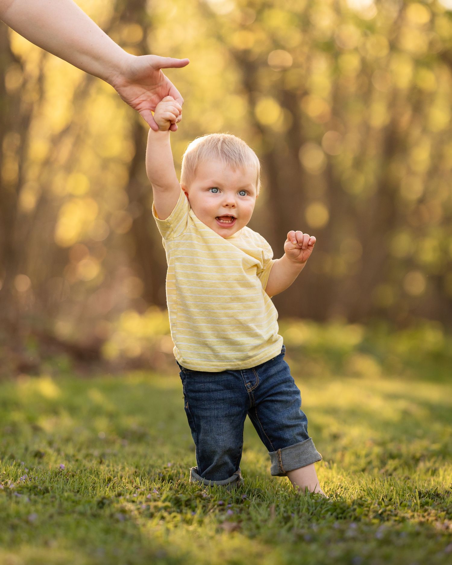 Smiling toddler boy in yellow shirt taking first steps holding an adult hand in golden light.
