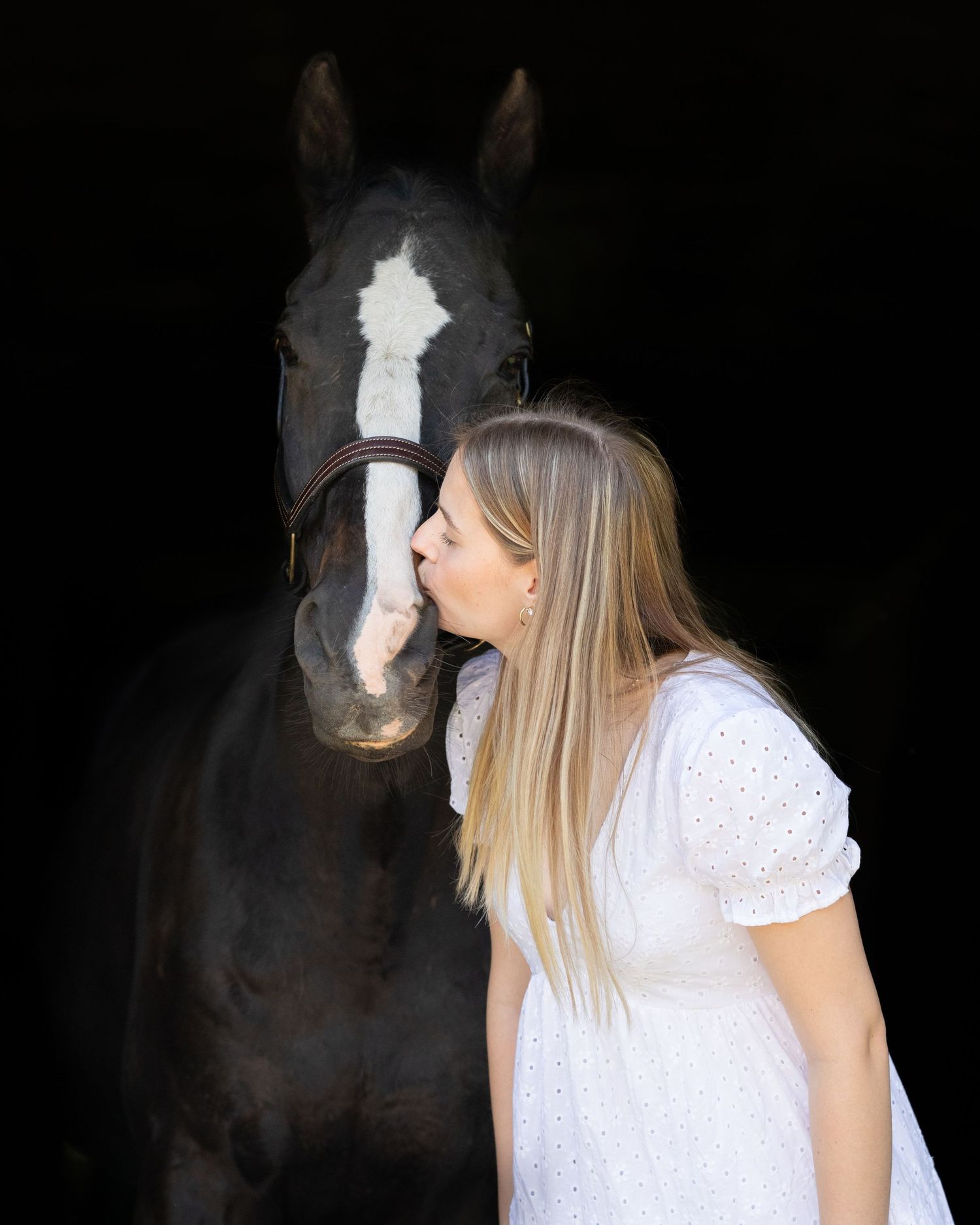Young woman in white dress kissing black horse on nose against dark background.