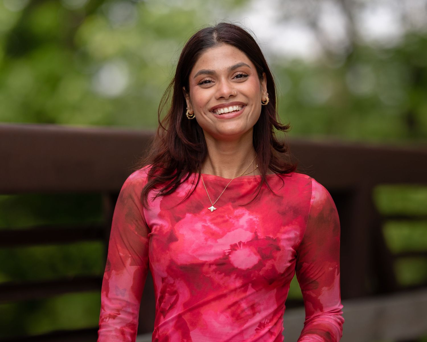 Smiling woman in pink floral mesh dress standing on a wooden bridge with green trees behind her.