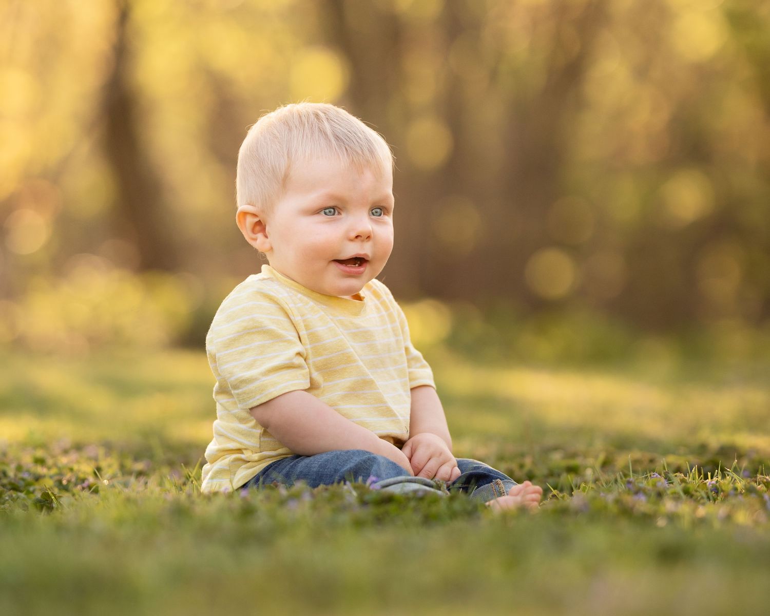 Happy baby boy in yellow shirt sitting on green grass smiling in a golden sunlit park.
