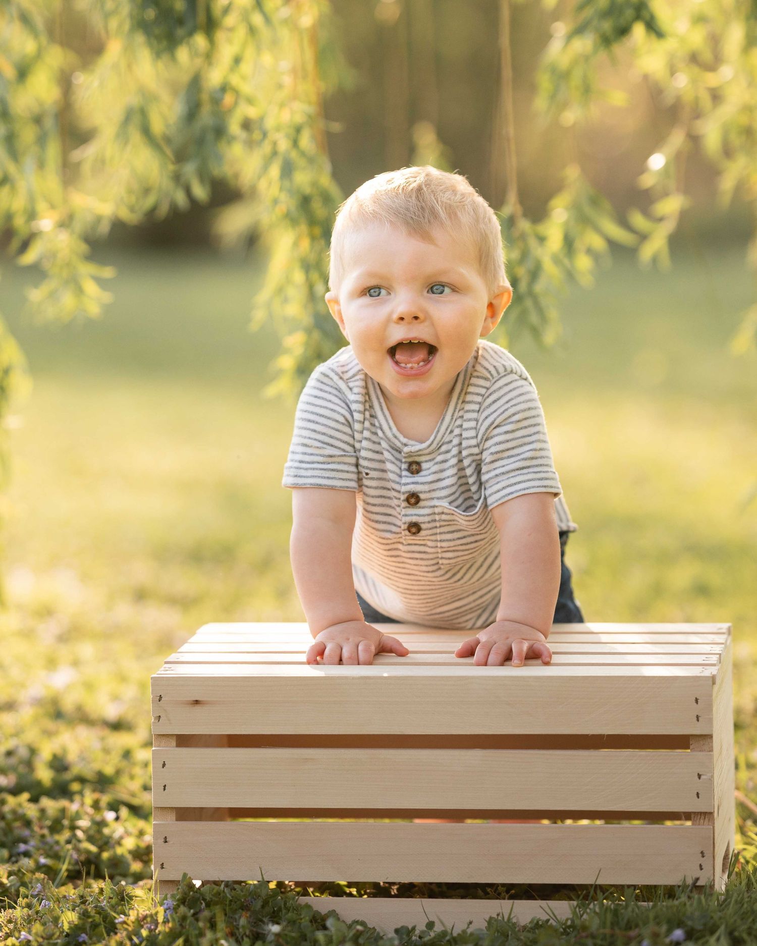Happy toddler boy leaning on a wooden crate outdoors in golden sunlight with a big smile.
