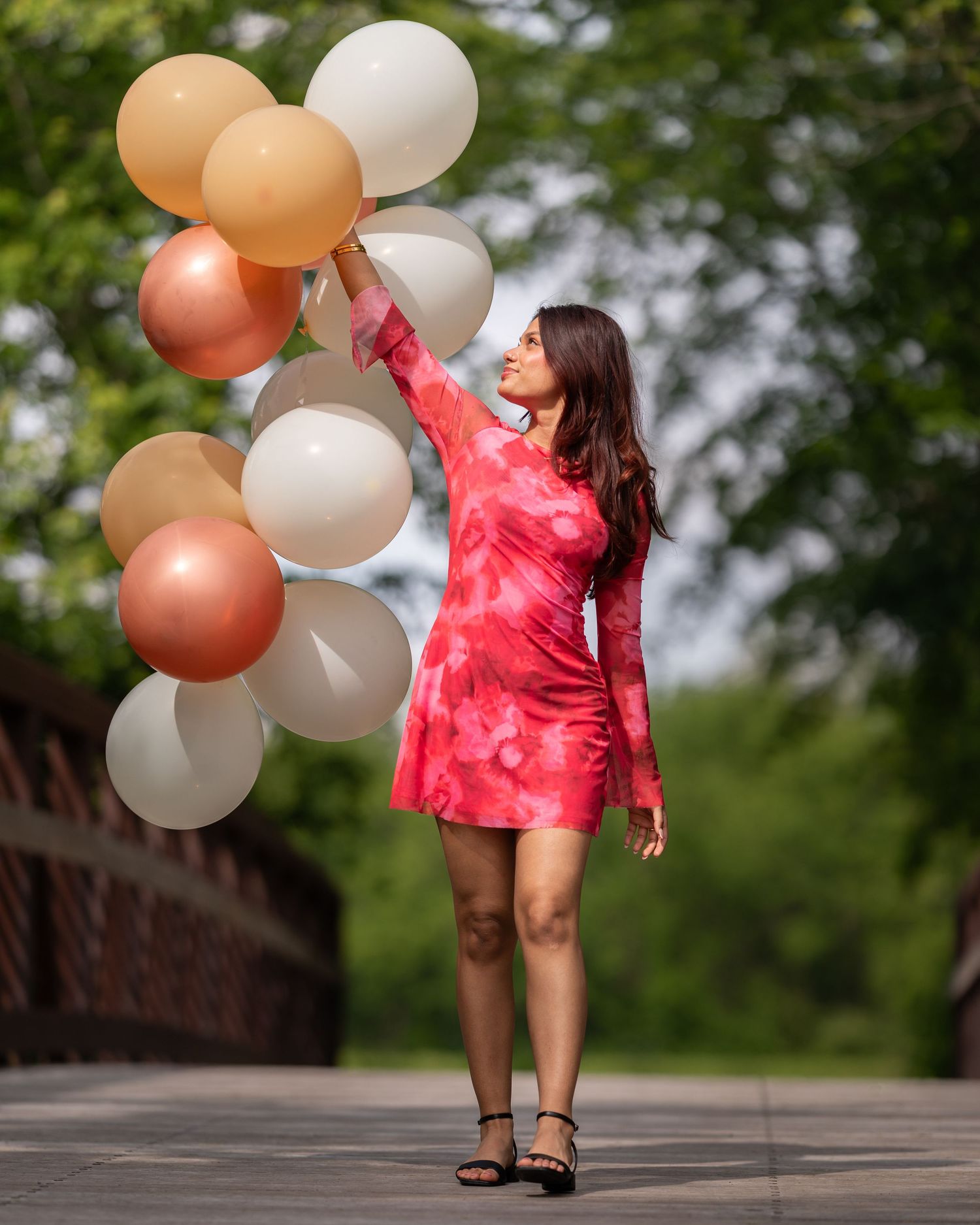 Woman in red tie-dye dress holding colorful balloons outdoors on a bridge surrounded by green trees.
