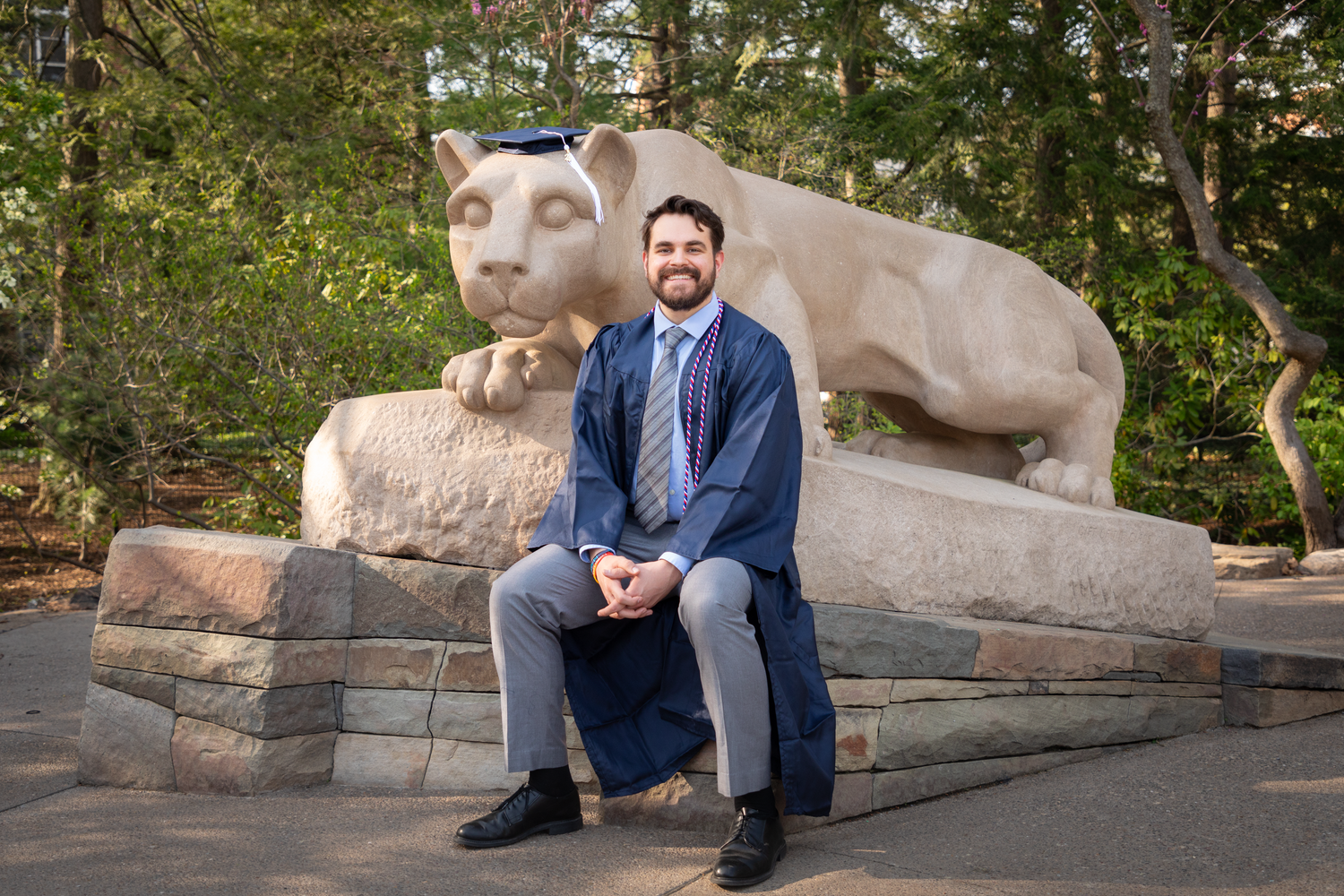 Man in suit sitting before Penn State Nittany Lion shrine for graduation portrait outdoors.