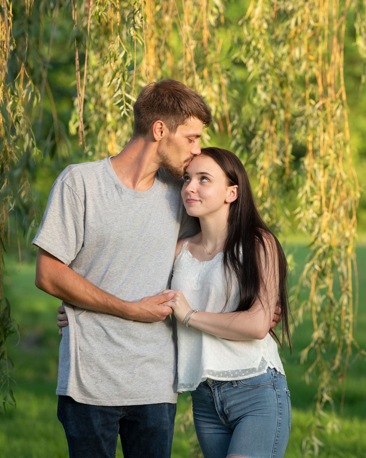 Young couple sharing tender moment under weeping willow tree in golden sunlight.