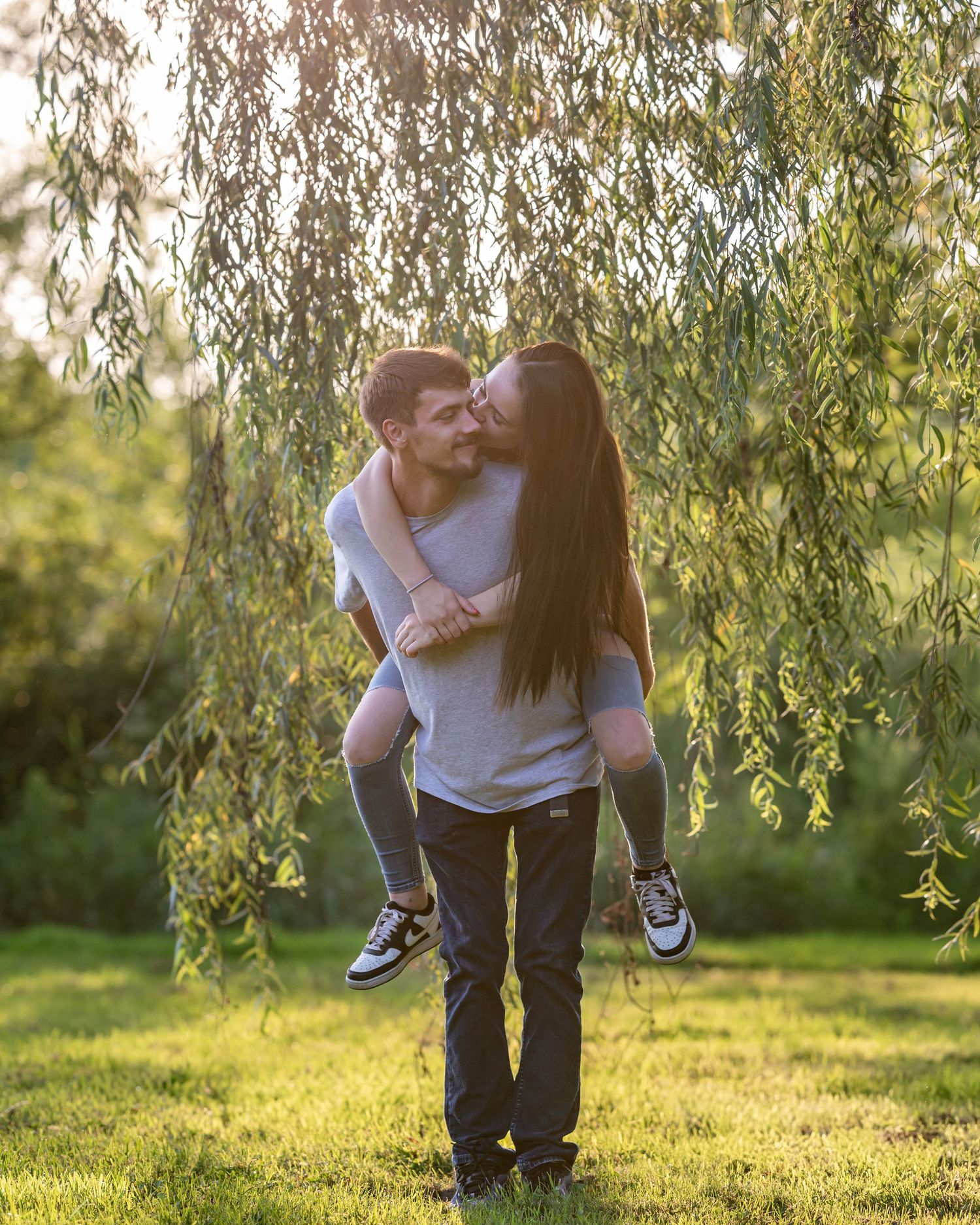 Man giving woman piggyback ride while kissing under willow tree in sunny park.
