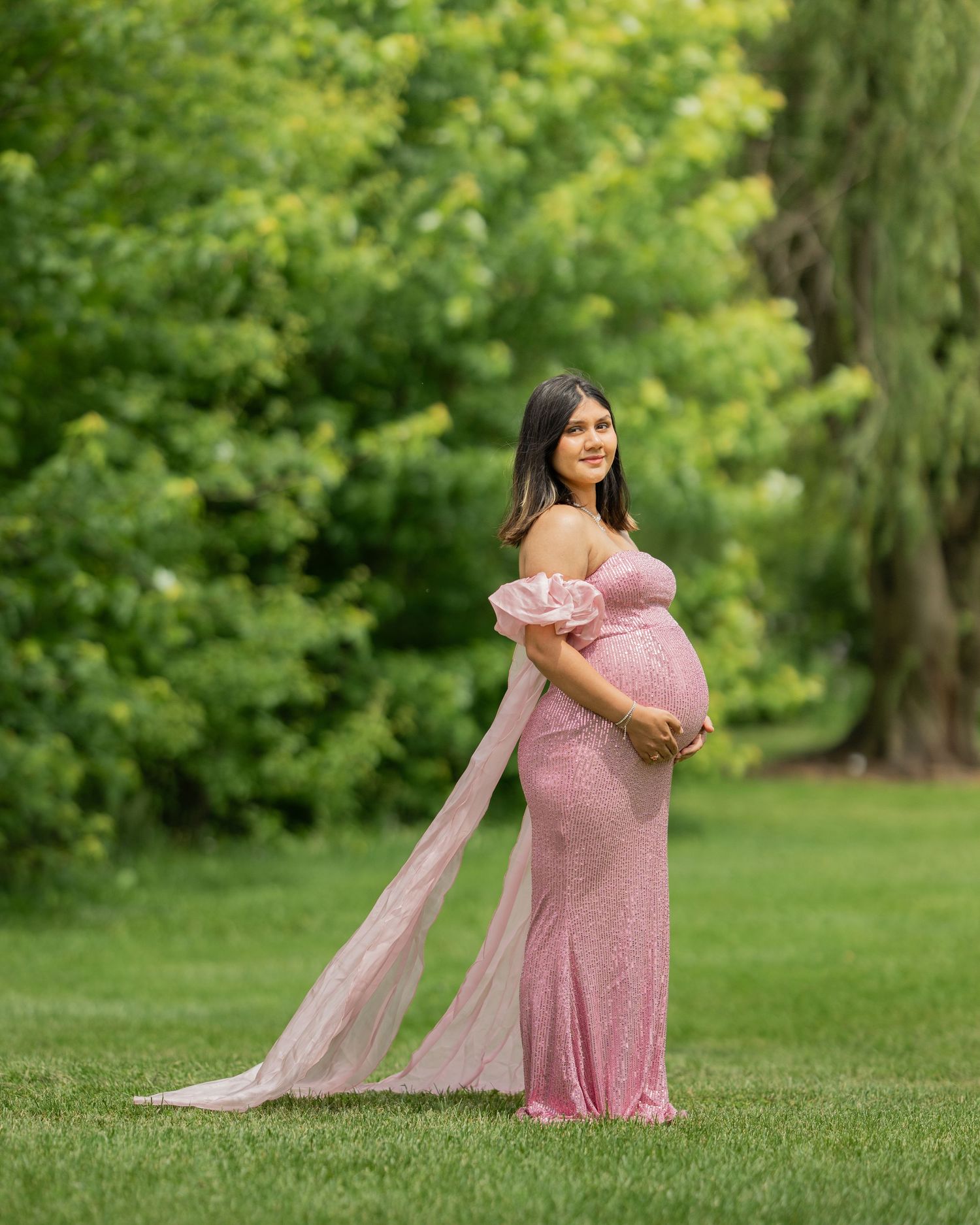 Pregnant woman in elegant pink sequin maternity gown posing outdoors in a green park setting.
