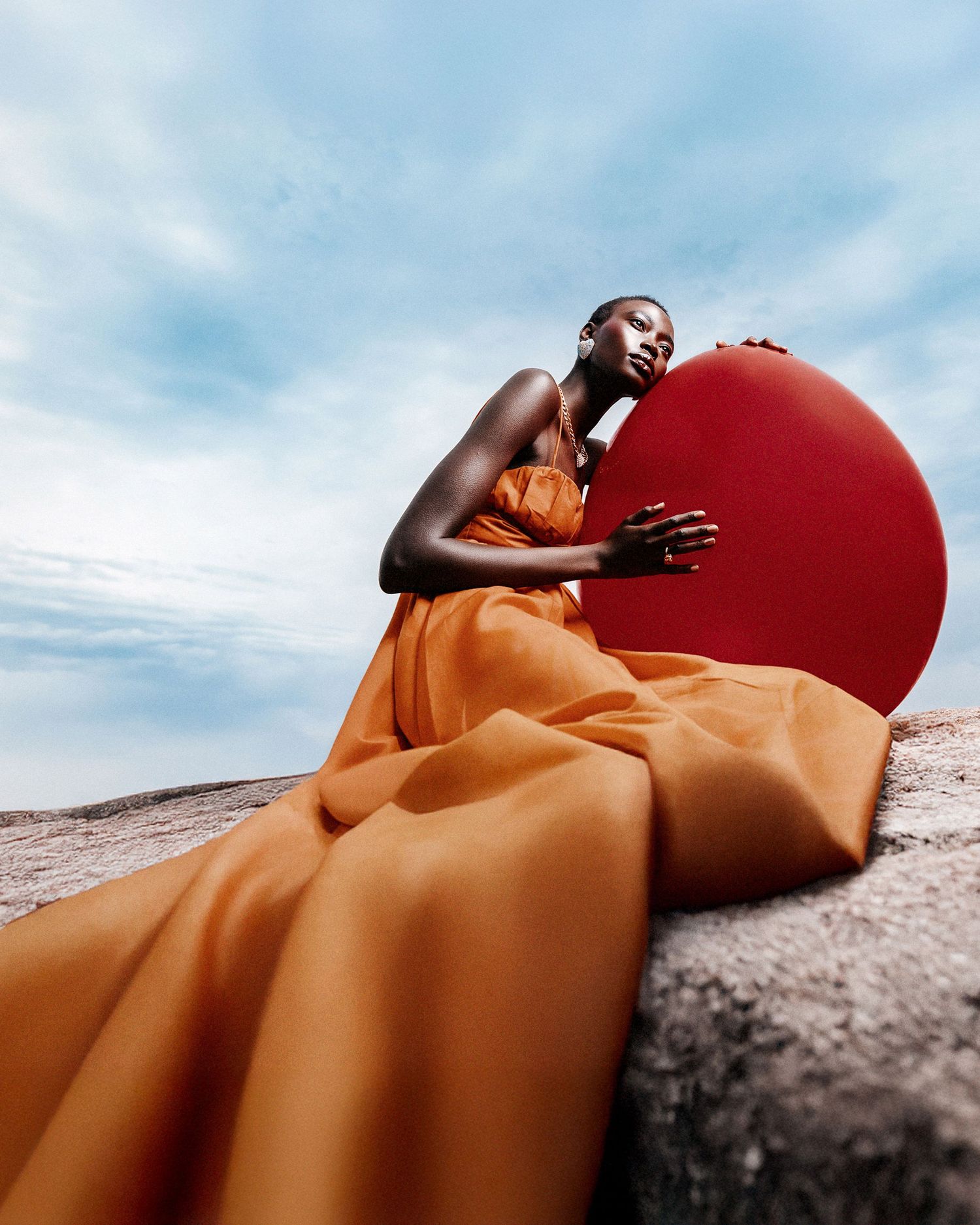 Artistic portrait with flowing orange fabric and large red sphere against dramatic sky.