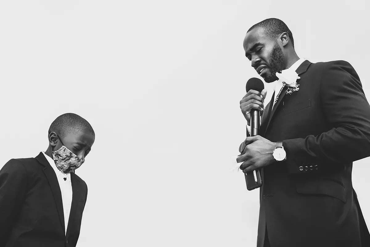 Two people in formal black suits share a moment during a speech at an elegant event.