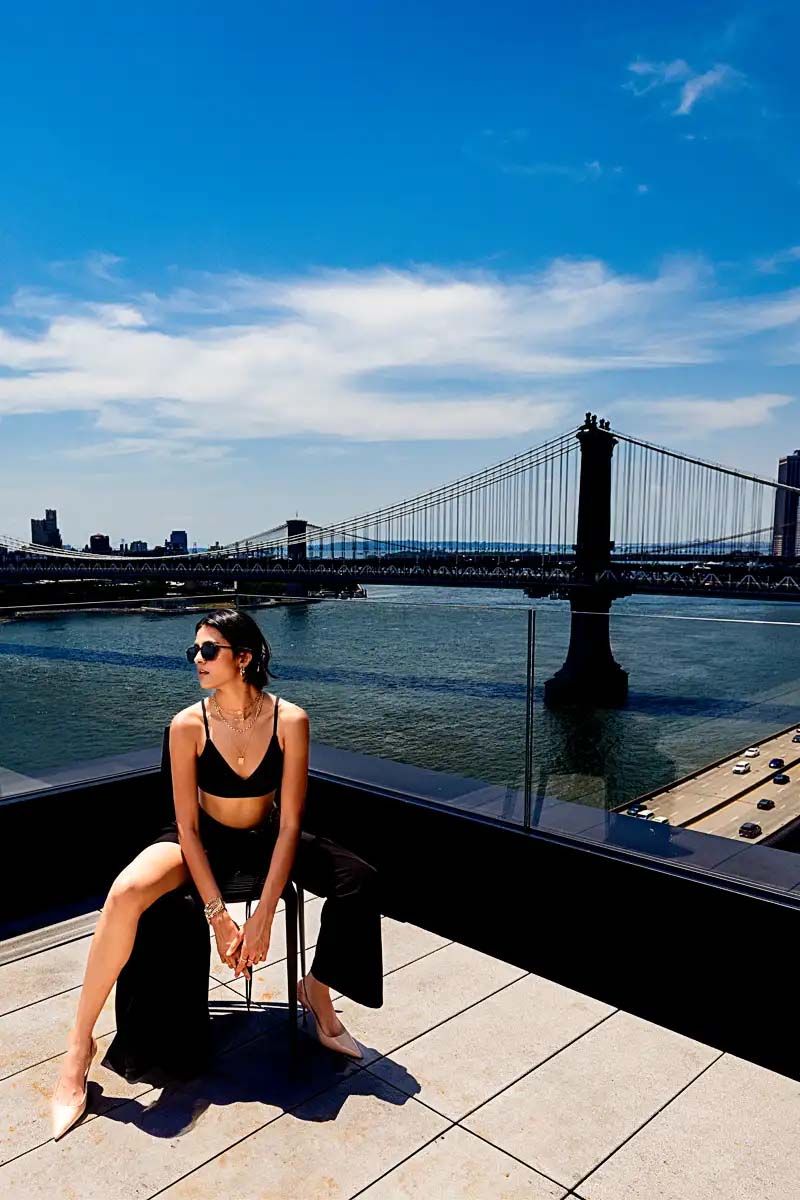 A rooftop photo with Manhattan Bridge in background showing person in black outfit sitting on ledge.